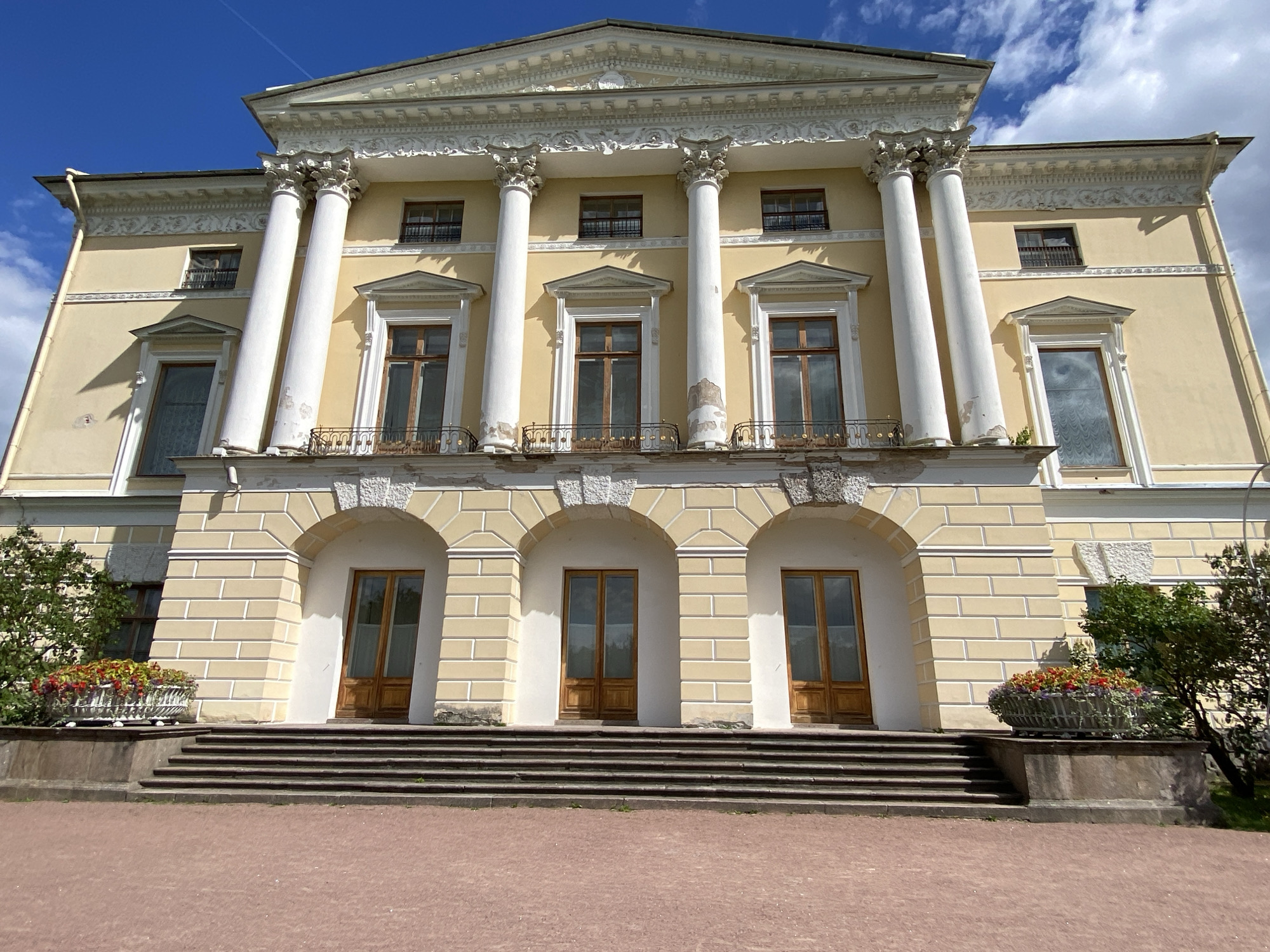 The image shows the facade of a classical-style palace, likely part of the Pavlovsk State Museum-Reserve in Pavlovsk, Russia, under a partly cloudy blue sky. The palace is painted a pale yellow and features a prominent portico with four large, white, fluted columns supporting an ornate entablature. Above the columns, there is a pediment decorated with relief sculptures. The building has several tall, rectangular windows with wooden frames on the upper level, some with wrought-iron balconies. Below the portico, there are three arched entrances, each with a double wooden door. A set of wide stone stairs leads up to these entrances. In the foreground, a reddish-brown path is visible, and on either side of the stairs, there are large planters filled with colorful flowers. Green trees and shrubs are present to the left and right of the building. The architecture suggests a grand, historical estate.