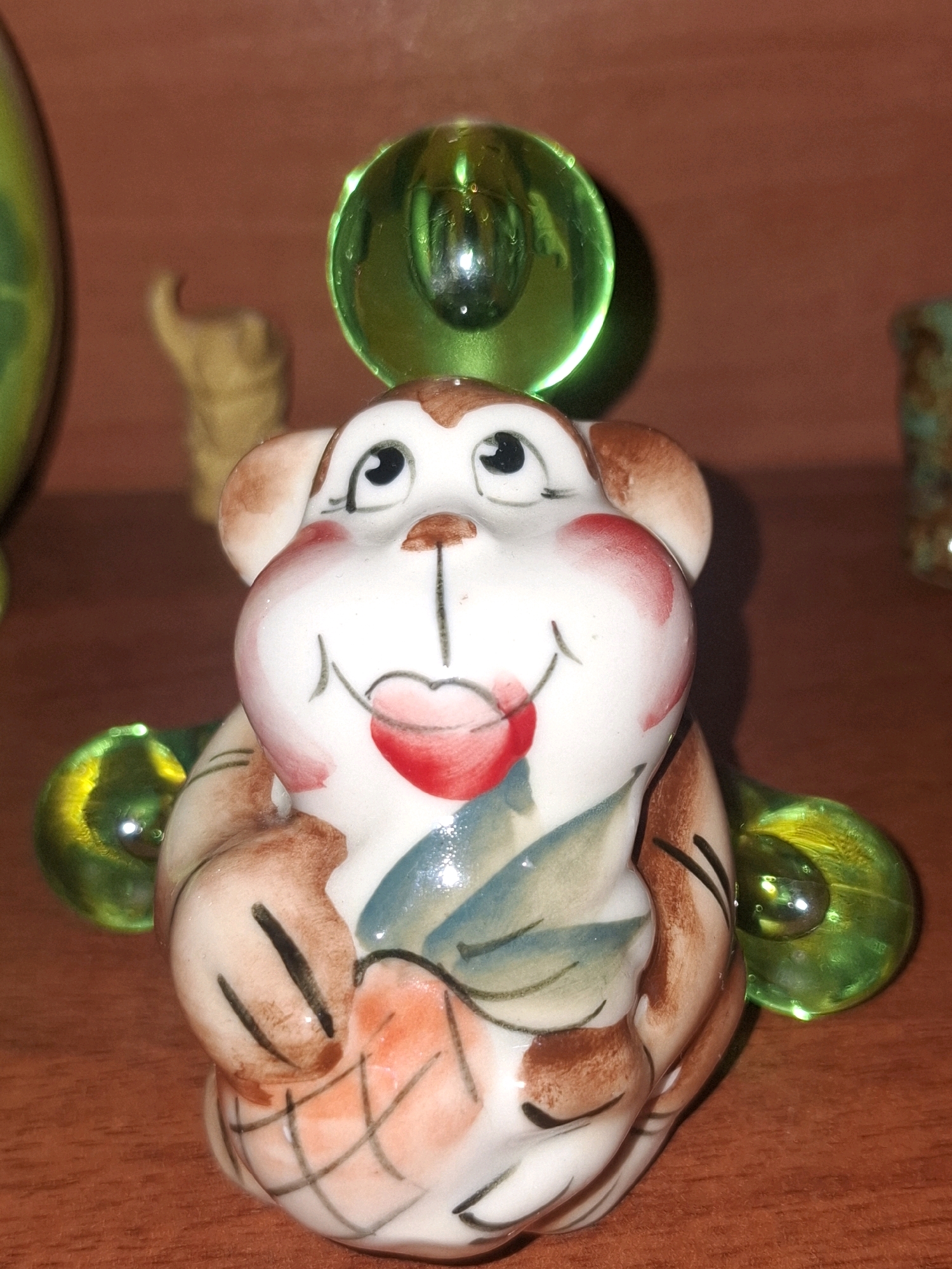 A close-up, eye-level shot shows a small, ceramic monkey figurine. The figurine is primarily white with brown and pink accents, depicting its face, ears, and arms. It has black eyes and a red, heart-shaped mouth. The monkey is holding a reddish-orange object, possibly fruit, with a grid pattern painted on it. Two green glass orbs are attached to the sides of the figurine, one on each arm, with visible reflections. The figurine is placed on a wooden surface, possibly a table, with a blurred brown background. There are no people or specific locations in Almaty, Kazakhstan visible in the image.