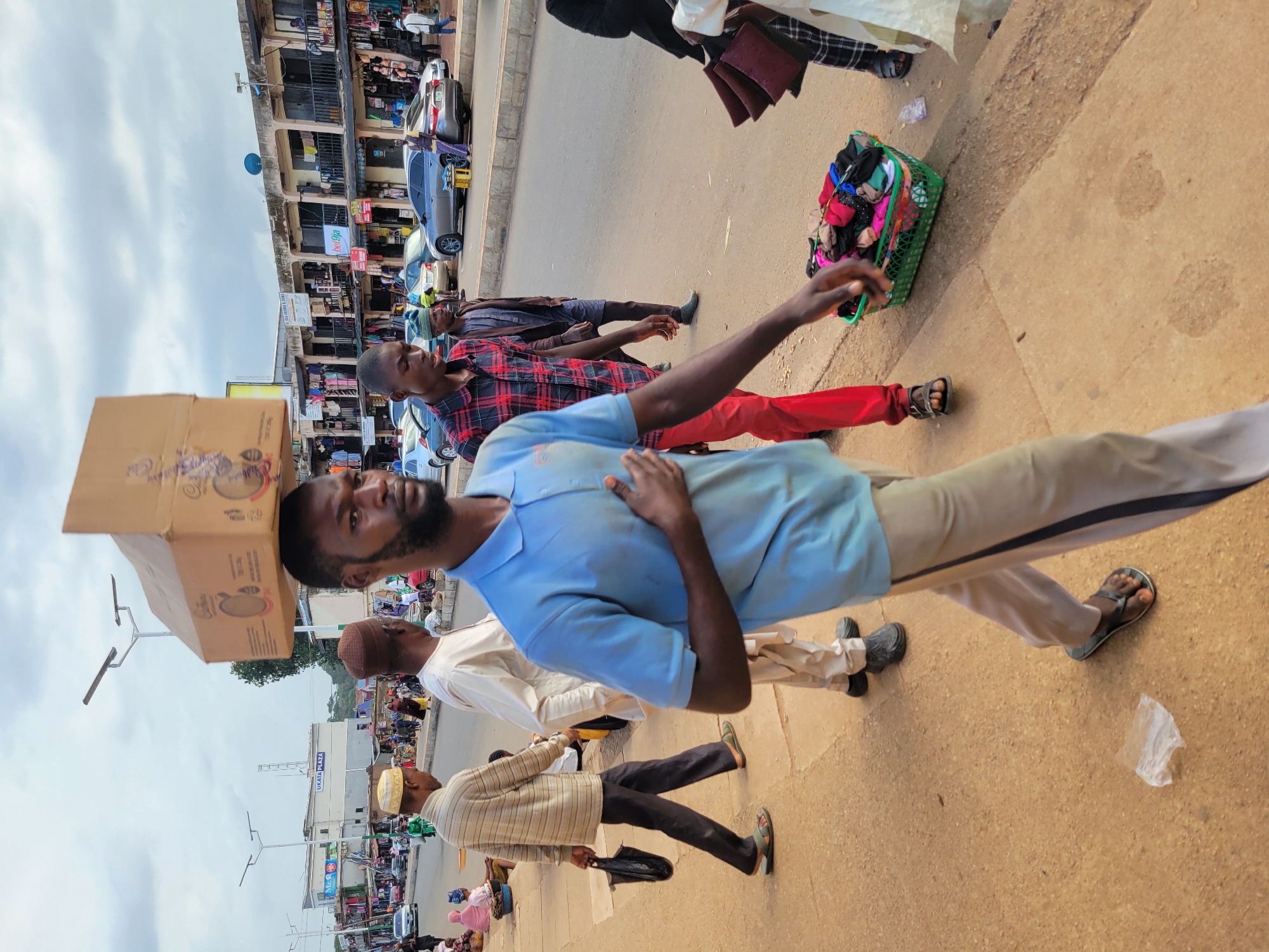 This media file depicts a busy street scene, likely a market, with several people and objects. The primary subject is a man carrying a large cardboard box on his head, wearing a light blue short-sleeved shirt and beige pants. He is walking towards the viewer. To his right, another man in a red and black plaid shirt and red pants stands, with a green basket filled with colorful clothing items next to him. Further back, other individuals can be seen walking and interacting. The background reveals shops with multiple stories, cars parked along the street, and streetlights. The ground is paved with concrete and sand.
The image appears to be taken during the daytime, with overcast skies suggesting potentially cloudy weather. The overall atmosphere is one of activity and commerce. There are no strong emotional cues visible, but the scene suggests a bustling daily routine.
Visible text includes "UKATA PLAZA" on a building in the background. Other signs are present but illegible.
