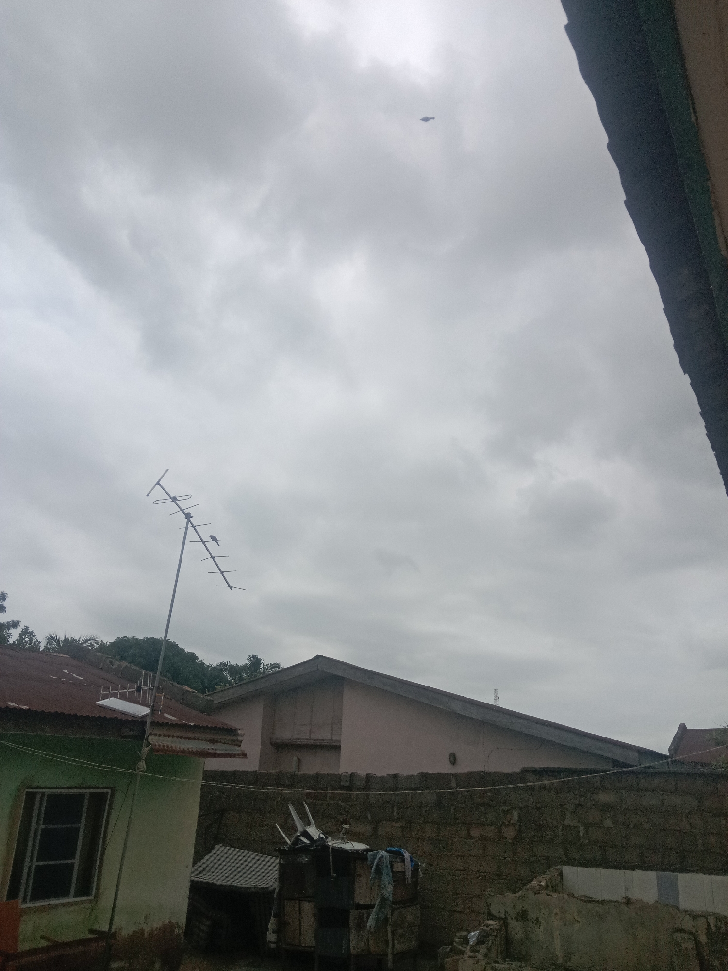 A dull, overcast day is depicted in this image, taken from a rooftop perspective in Lokoja, Nigeria. The sky is filled with heavy grey clouds. In the middle ground, a television antenna stands tall, angled against the sky, with two birds perched on its structure. Beyond the antenna, a pale pink building with a triangular roofline is visible, partially obscured by a rough-hewn stone wall in the foreground. To the left of the pink building, a green house with a rusted corrugated iron roof is partially visible, featuring a window with metal bars. Several small structures and debris are scattered around the foreground, suggesting a residential area that may be undergoing some disrepair or construction. In the far distance, a small dark object, possibly a drone or a bird in flight, is visible against the cloudy sky.