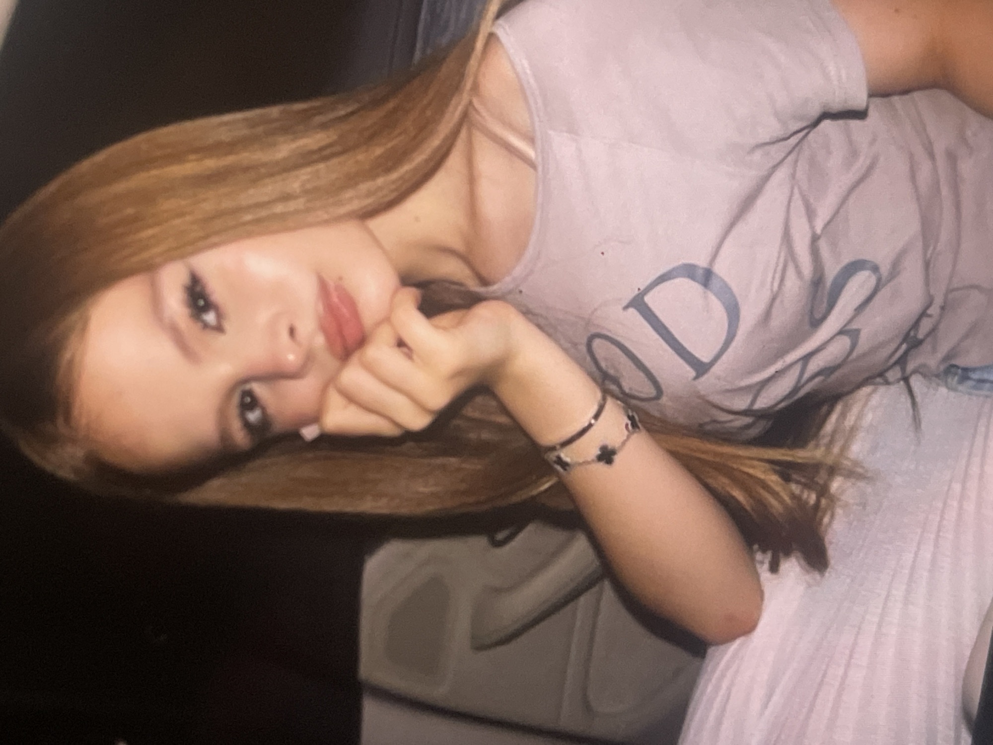 An image, rotated 90 degrees clockwise, captures a young woman posing in an indoor setting. She has long, light brown hair that cascades over her shoulder and arm. Her face, framed by her hair, is turned towards the camera, and she props her chin or jaw with her right hand, showcasing manicured, light-colored fingernails.
She wears dark eyeliner and mascara, emphasizing her eyes, and a glossy pink or reddish lipstick on her full lips. Her expression is calm and direct, with a hint of thoughtfulness.
She is dressed in a light-colored (possibly light grey or beige) short-sleeved t-shirt. Visible on the t-shirt are large, dark letters that clearly spell "ODS", likely part of a larger word such as "GOODS" or "GODS", with the initial letter mostly obscured. On her left wrist, she wears two bracelets: a thin, black, rigid bangle and a chain bracelet adorned with multiple small, black, four-leaf clover-shaped charms.
The background is softly blurred, showing dark areas and some lighter, textured surfaces, suggesting she is reclining against furniture or perhaps a car seat. The lighting is soft and even, typical of an indoor environment, making it difficult to discern the time of day. There are no visual cues within the image itself to specifically indicate the provided location context of Poltava, Ukraine.