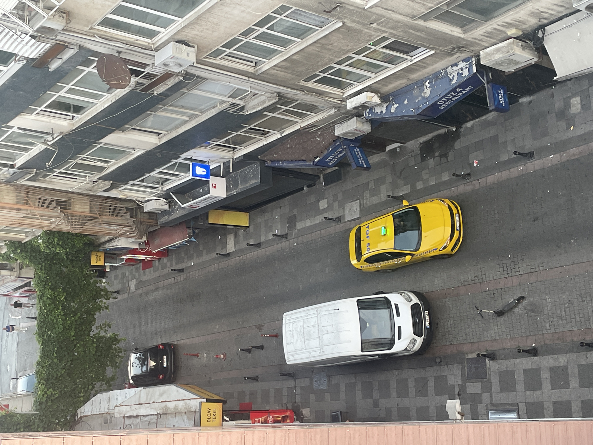 An aerial, high-angle shot captures a narrow street in Kadıköy, Turkey, lined with buildings and parked vehicles. Two people, a male and a female, walk away from the camera on a sidewalk bordered by lush green foliage, with a dark-colored car parked on the street. Further down the street, a white van is parked, and beside it, a yellow taxi is stopped. A dark-colored electric scooter lies on its side on the pavement. The buildings on the right are multi-story, featuring numerous windows and balconies, with some displaying signage for "OTUZ 4 RESTAURANT." The street appears to be paved with cobblestones and asphalt.