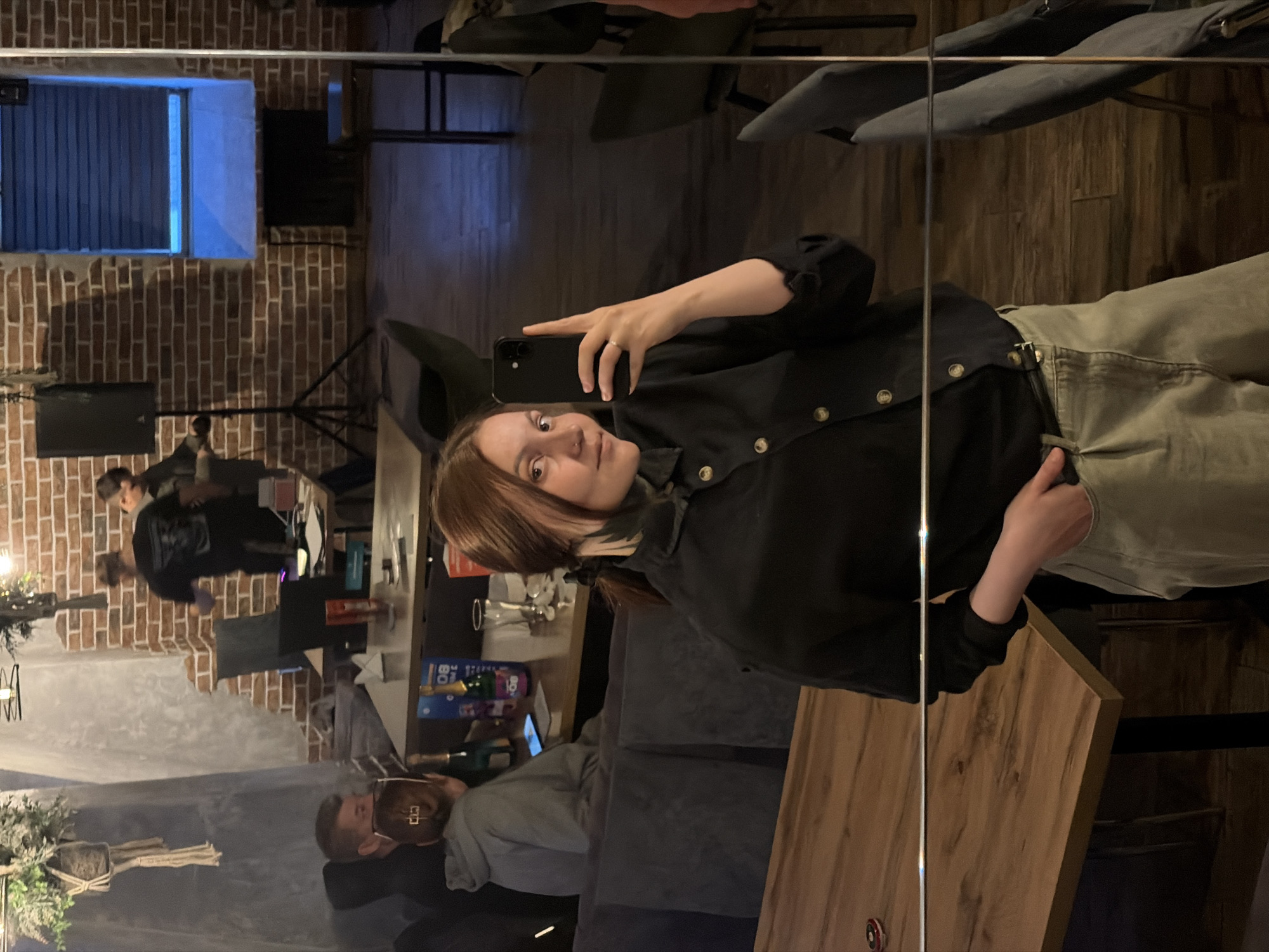 The image is a mirror selfie taken indoors, capturing a young woman and the reflection of a cafe, bar, or restaurant interior in Chaikovsky, Russia. The setting features exposed red brick walls, wooden tables, and dark seating, creating a rustic and dim atmosphere, suggesting evening or night.
The main subject is a young woman with shoulder-length brown hair, possibly with lighter streaks. She is wearing a dark, possibly black, button-up shirt or jacket with gold-colored buttons and light-colored pants, perhaps olive green. She is holding a black smartphone to take the picture, and a simple ring is visible on her left ring finger. Her expression is calm with a slight smile.
In the reflection, several other people are visible. On the left, two figures are partially seen, and further down, a man with a beard and glasses is seated at a table. Another person with brown hair pulled back is also visible. The reflected background includes a window with vertical blinds that glows with a blue light, dark speakers or rectangular objects, and decorative potted plants. On one of the tables, a blue and white box with the number "80" printed on it is visible, along with dark bottles, possibly champagne. The mirror itself has a vertical dividing line running through the image.