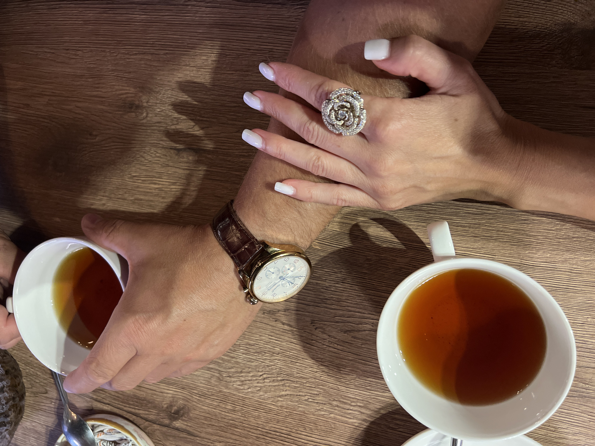 The image shows a close-up view of two people's hands resting on a wooden table, with two white teacups filled with dark liquid, likely tea, positioned on the table. One person's hand, appearing to be male, is wearing a gold watch with a brown leather strap and is holding one of the teacups. The other person's hand, appearing to be female, is adorned with a large, ornate ring featuring multiple small stones, possibly diamonds, in a floral design. The nails of this hand are painted white. The environment is a dimly lit indoor setting with a visible shadow cast by the hands onto the textured wooden surface of the table. No specific details indicate a location in Sochi, Russia.