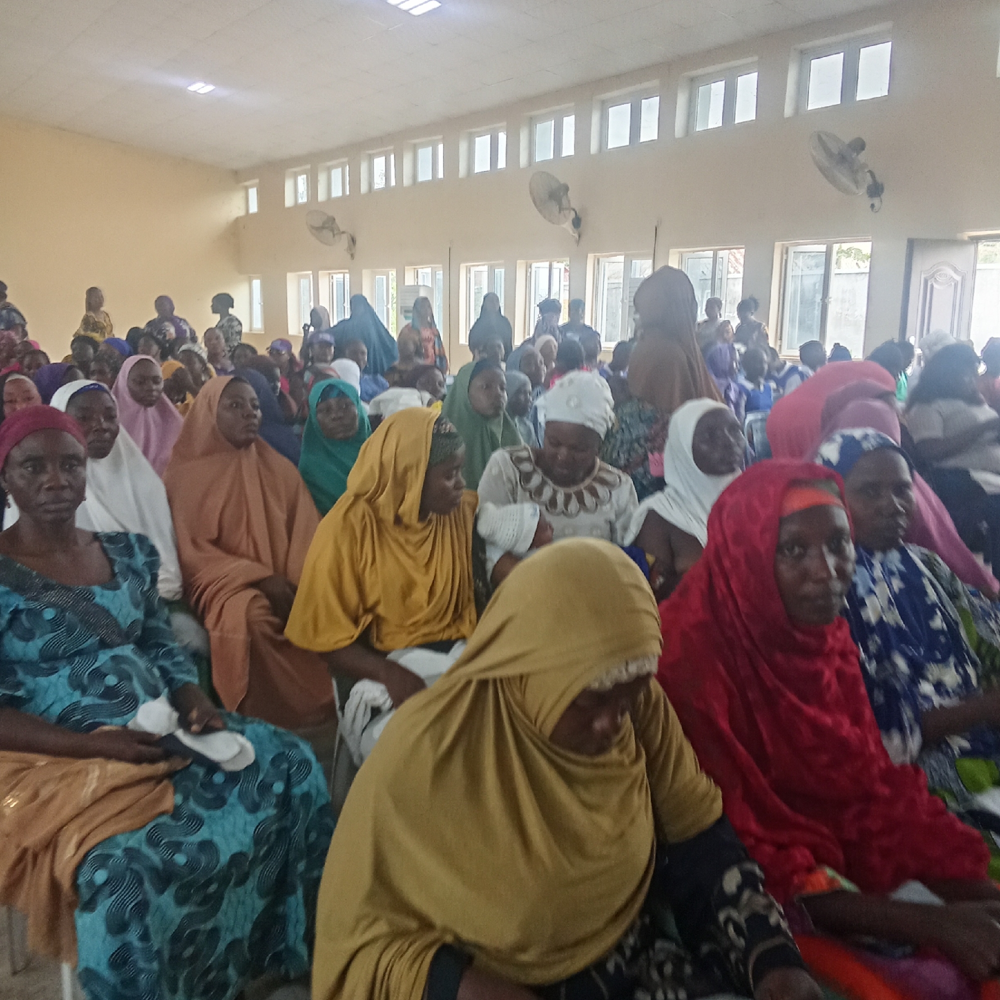 The image shows a large group of women seated in a room in Akwanga, Nigeria. The women are dressed in various colorful traditional attire, many wearing headscarves or hijabs of different colors including yellow, red, white, brown, and green. They are seated in rows, facing the same direction. The room has multiple rows of windows along the top of the walls and electric fans hanging from the ceiling. The walls are light-colored. The lighting is bright.