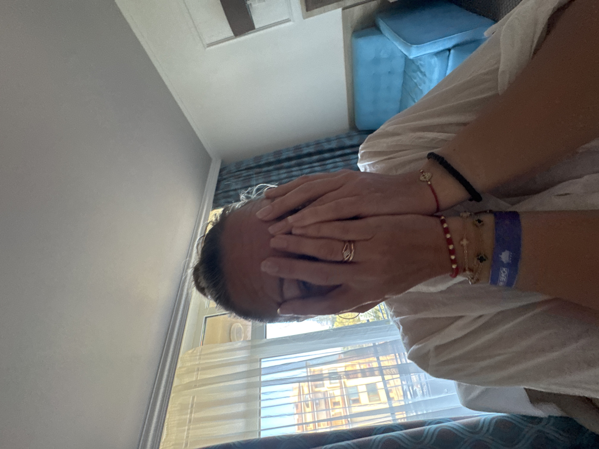 A woman, her face obscured by her hands, is seated in a room with a window revealing a building in Nesebar, Bulgaria. She wears a white garment and several bracelets on her wrists, including a blue wristband that reads "BEACH". A blue chair is visible in the background.