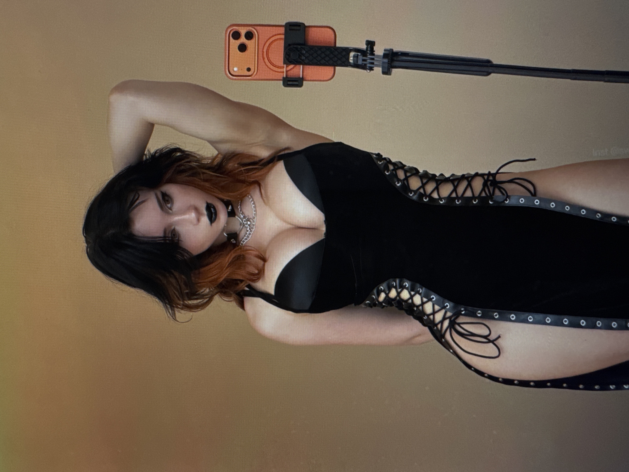 A young woman with dark hair with auburn highlights is posing for a photo with her right hand behind her head and her left arm down. She is wearing a black dress with a plunging neckline and a thigh-high slit on the left side, adorned with grommets and lacing. She has on a silver chain necklace and dark lipstick. An orange smartphone on a tripod is visible to her right. The background is a plain, muted gold color.
