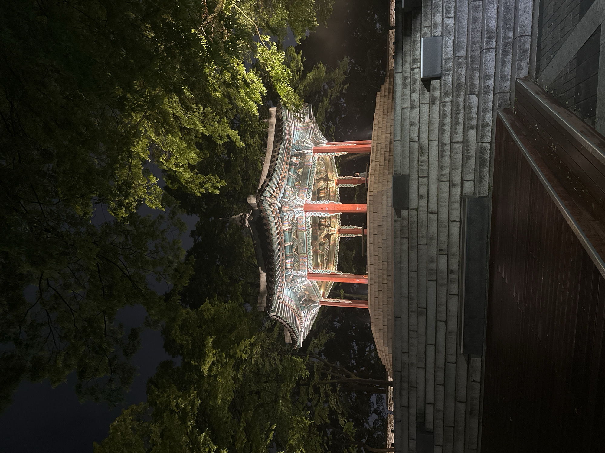 The image captures a traditional Korean pavilion illuminated at night, nestled among lush green trees. The intricate details of the pavilion's roof and red pillars are visible under the lighting. Adjacent to the pavilion is a stone staircase with a wooden railing. The setting is likely within the city of Seoul, South Korea, given the traditional architecture. The image lacks the presence of people.