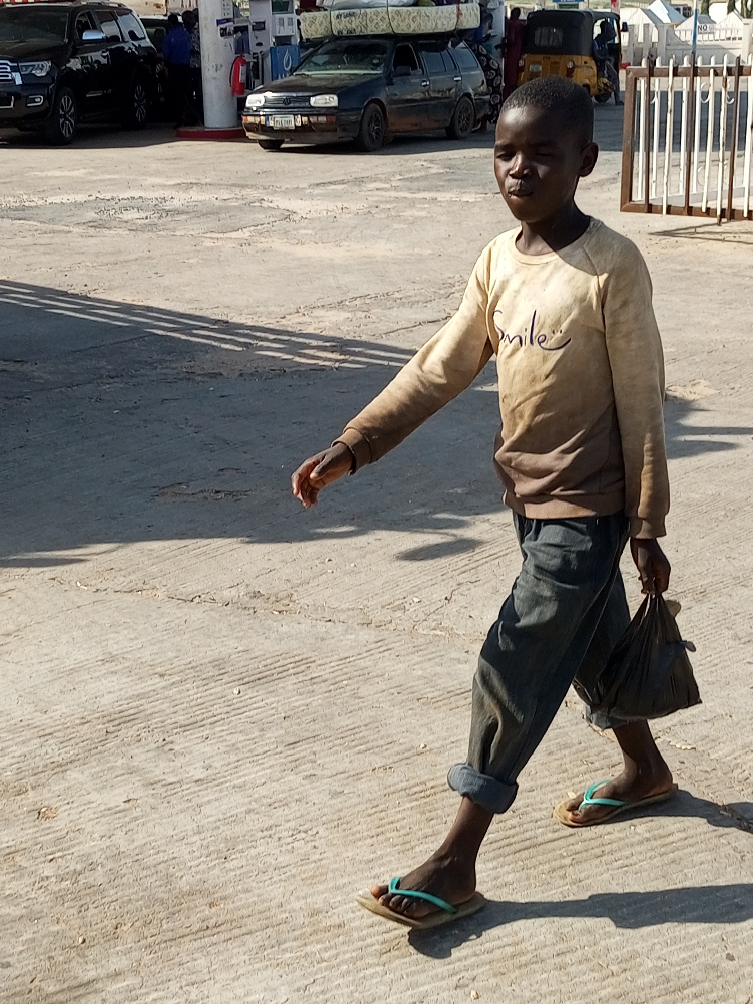 A young Black boy walks across a sunlit, dusty concrete surface in Gombe, Nigeria. He is wearing a faded, light-colored long-sleeved shirt with "Smile" written on it in blue, cuffed blue jeans, and blue flip-flops. The boy is holding a black plastic bag in his right hand. In the background, there are several vehicles, including a black SUV, a dark sedan, and a yellow auto-rickshaw. A gas station with pumps and a canopy is also visible. The ground is worn and textured, with tire tracks and debris. The overall scene suggests a common street or roadside environment within the city.