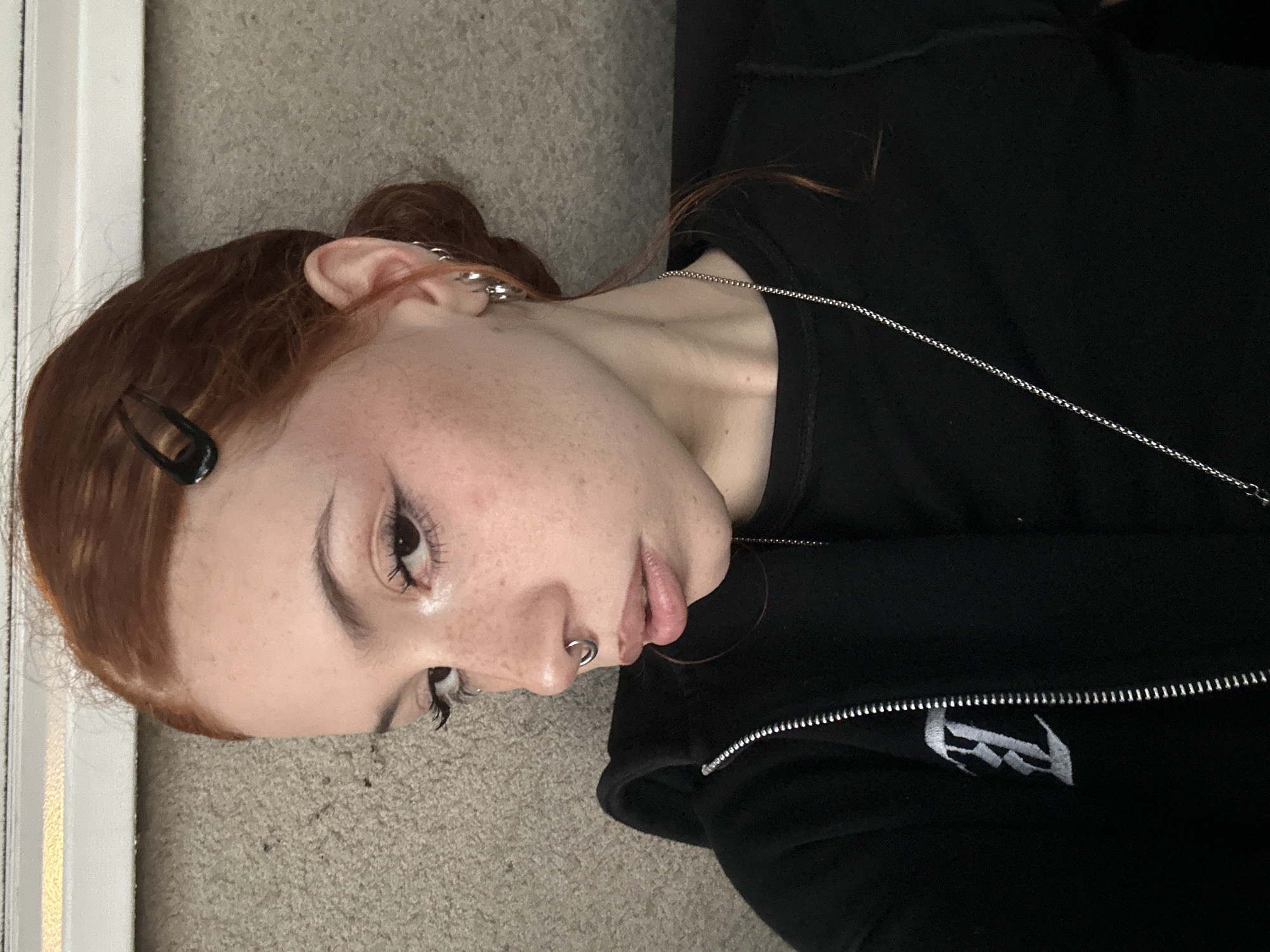 A young individual with reddish-brown hair is depicted lying on a light grey carpeted floor, positioned next to a white vertical structure that appears to be a door frame or wall. The subject has fair skin with numerous visible freckles across their nose and cheeks. Their hair is partially styled back, secured on one side by a black hair clip.
The person's face is shown in profile, looking slightly upwards and to the right with a neutral to contemplative expression. Notable facial features include dark eyebrows and eyelashes, accentuated by what appears to be eye makeup. A silver septum piercing, in the form of a ring, is visible on their nose. Their left ear is adorned with multiple silver hoop and stud piercings. Around their neck, a thin silver chain necklace is worn.
The individual is dressed in a black zippered hoodie. On the left side of the chest, there is a distinct white embroidered logo, stylized to resemble a gothic 'R' or similar decorative initial. The overall scene suggests an indoor setting. This media is contextualized as being from Buenos Aires, Argentina.