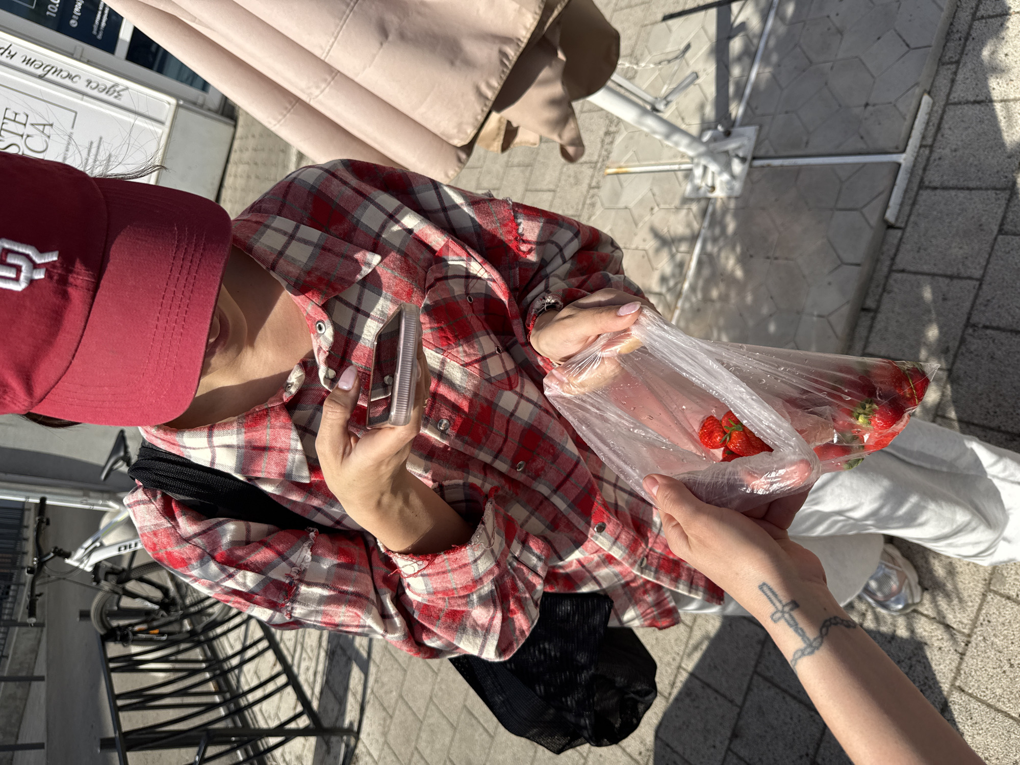 A top-down shot shows two individuals interacting on a sunny outdoor day. One individual, wearing a red and white checkered long-sleeved shirt and a red baseball cap, holds a plastic bag containing strawberries. The person also holds a metallic object, possibly a phone or wallet, in the other hand. Another person is extending their hand to receive the strawberries. The person's arm is visible, and a tattoo of a cross and chain is seen on their forearm.
A black mesh bag is on the first person's shoulder. The background includes a bicycle rack with bicycles, a tan umbrella, and paving stones. The exact location in Krasnoyarsk is not determinable from this image alone.