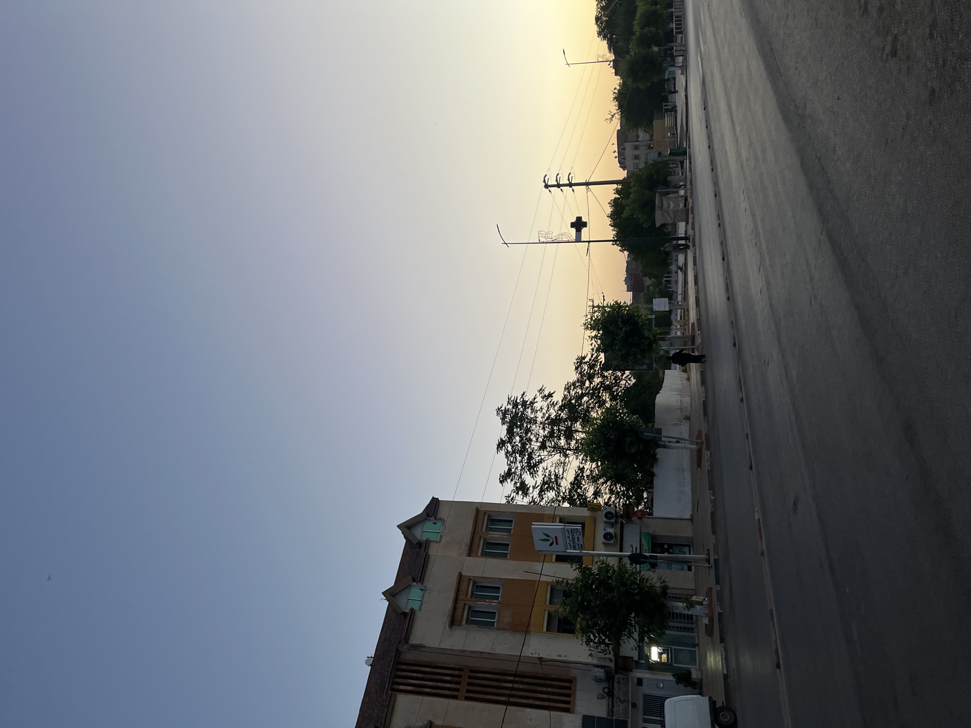 The image depicts a street scene in El Hajeb, Morocco, during twilight. On the left side of the street, a multi-story building with a beige facade and distinctive gabled dormer windows stands. A sign with Arabic script and a green logo is mounted on a pole in front of the building, alongside an air conditioning unit. Trees line the sidewalk in front of the building, and a white van is parked to the far left. The street itself is paved, with a curb and what appears to be a pedestrian walkway. Several streetlights are visible, one of which has an ornate design at its top. In the distance, more trees and buildings can be seen, with power lines crisscrossing the sky. The sky transitions from a clear light blue at the top to a warm yellow-orange hue near the horizon, indicating either sunrise or sunset. A few small figures, likely pedestrians, are present on the sidewalk and crossing the street in the middle ground. One person is walking away from the camera on the right side of the street. The overall atmosphere is calm and suggests a quiet urban environment at the close of day or beginning of the morning.