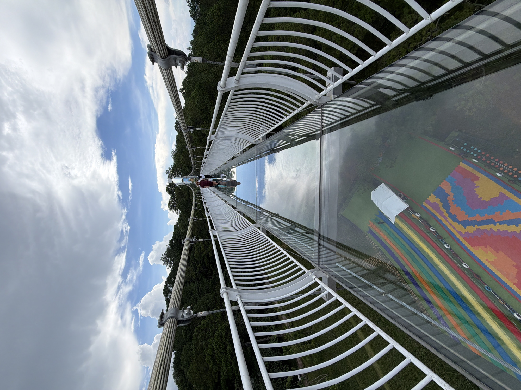 The image depicts a glass-bottomed suspension bridge in Lam Vien - Da Lat, Vietnam. The bridge is constructed with white metal railings and thick glass panels that allow a view of the forested area below. Several people are walking on the bridge, their reflections visible in the glass. The sky is filled with white, fluffy clouds against a blue backdrop, and lush green trees surround the bridge. The glass panels of the bridge also feature a colorful, geometric pattern beneath the surface, possibly a decorative element or part of the underlying structure.