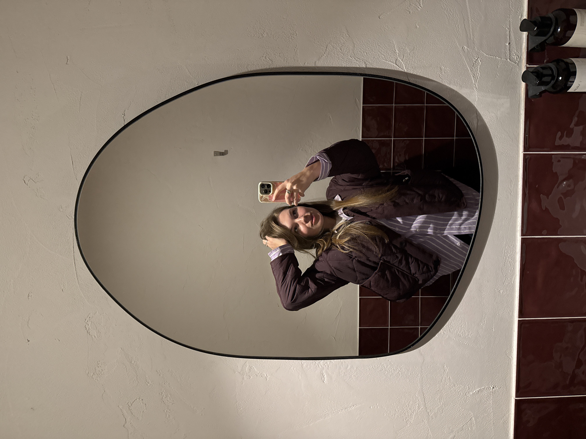 A young adult female is depicted taking a mirror selfie in an indoor setting, identified as located in Belgrade, Serbia. The subject, a female with long brown hair, is reflected in a large, irregularly shaped mirror with a thin black frame. She is wearing a dark purple quilted jacket over a light-colored, possibly striped, button-up shirt. Her right hand is raised, fingers lightly touching the hair on the top of her head, while her left hand holds a light-colored smartphone, visible in the reflection as she photographs herself. A ring is visible on her left ring finger. The wall behind the subject, also reflected in the mirror, features an upper section with a textured, off-white or light grey finish, and a lower section composed of glossy, dark red square tiles arranged in a grid pattern. A small, silver-colored wall hook is visible in the upper right quadrant of the mirror's reflection. Below the mirror, on a shelf or surface, two dark-colored dispenser bottles are mounted on individual black wall brackets, suggesting a public restroom or washroom environment.