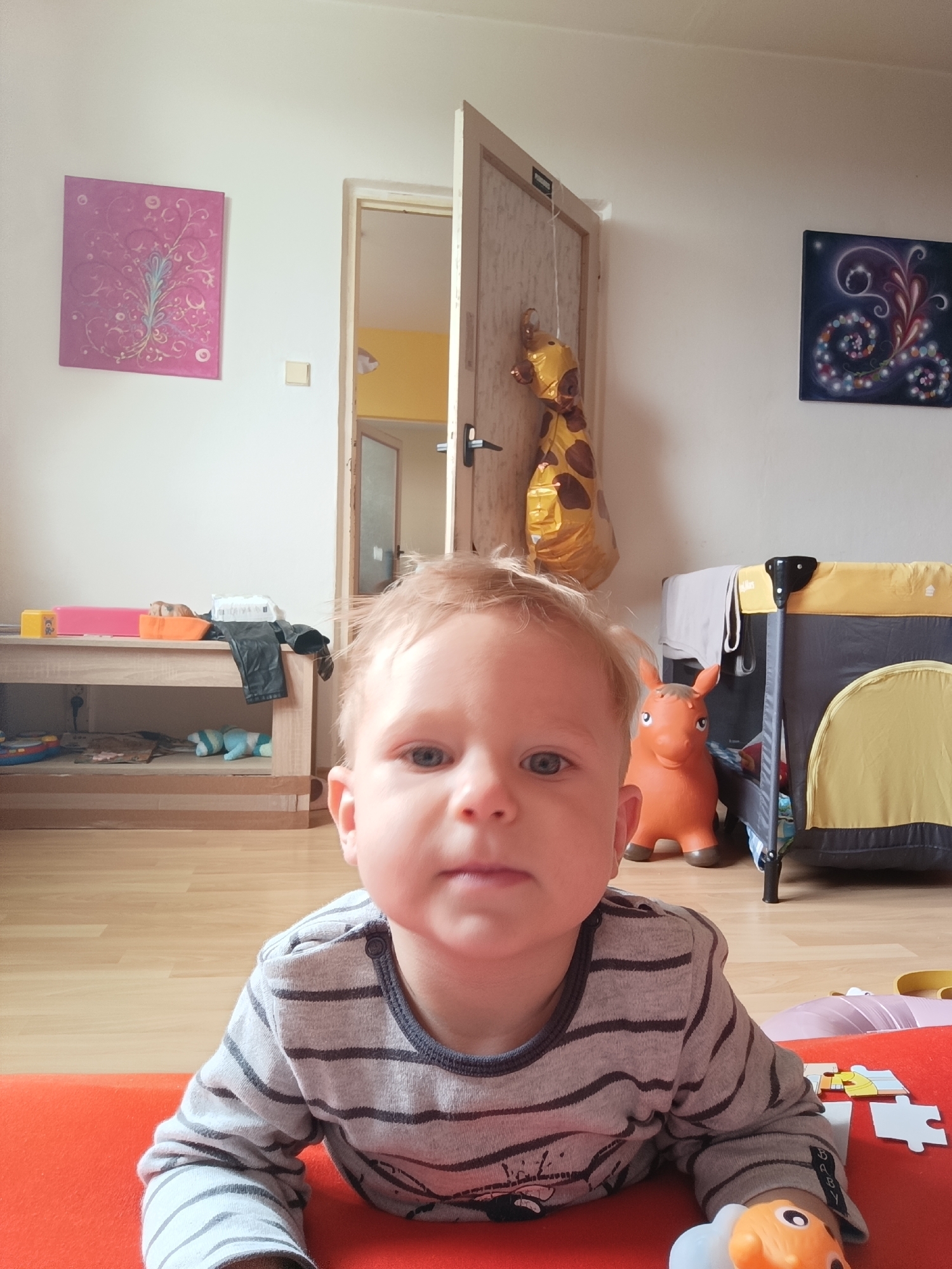 A young child with blonde hair and blue eyes is lying on an orange mat in a room. The child is wearing a gray and black striped long-sleeved shirt. In the background, there is a wooden table with various items on it, including toys and clothing. A baby crib with yellow and gray fabric is also visible. There is a wooden floor and white walls. Artwork, including a pink floral painting and a dark painting with swirling patterns, hangs on the walls. A giraffe-shaped balloon is tied to a door. An orange inflatable horse toy is placed on the floor. The scene appears to be in a residential setting, possibly a child's playroom or bedroom, located in Olomouc, Czechia.