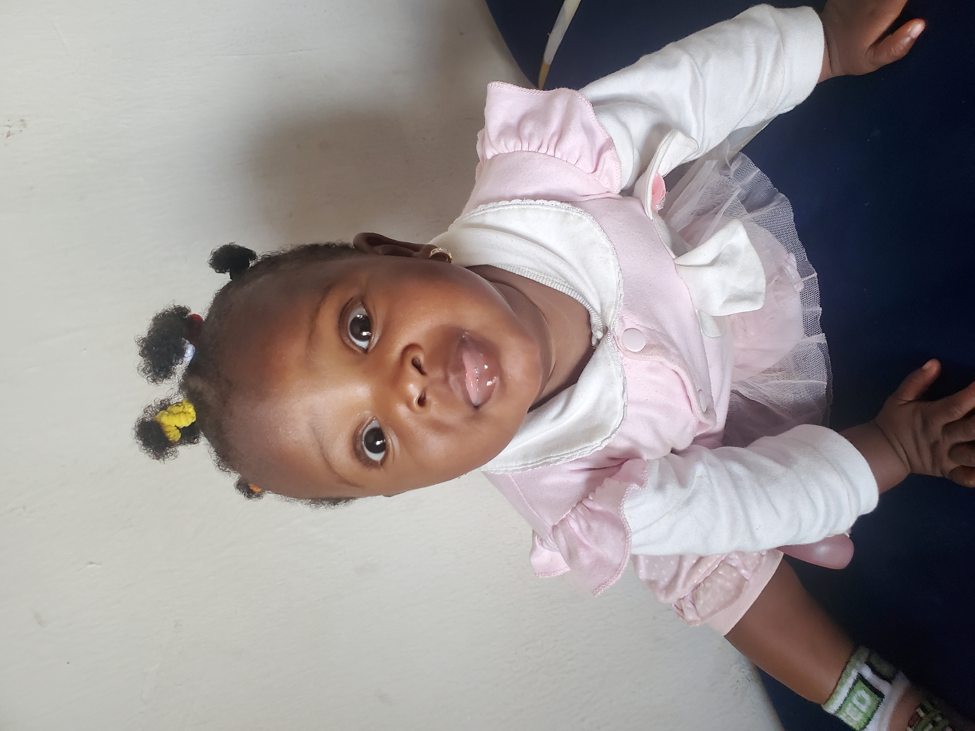 A close-up, eye-level shot captures a young Black female infant sitting on a dark blue surface against a plain, light-colored wall. The infant is positioned centrally in the frame, facing the camera directly. She has dark skin and her hair is styled in several small, dark pigtails adorned with colorful hair ties. The infant's eyes are wide open and directed at the camera, and her tongue is protruding from her mouth, giving her a playful or surprised expression. She is wearing a pink and white outfit. The pink part of the dress features ruffled sleeves and a skirt with a sheer, white tulle overlay. The white inner layer has long sleeves and a collar, and the pink outer layer has a button closure on the front. On her left arm, she wears a decorative bracelet with green and white patterns. Her hands are visible, one resting on the dark blue surface in front of her, and the other near her left leg. The background is a plain, off-white wall with some minor texture and faint discoloration, suggesting an indoor setting, possibly a home. The overall lighting appears to be natural, coming from the front. The specific location in Jos, Nigeria, is not discernible from the visual information in the image.
