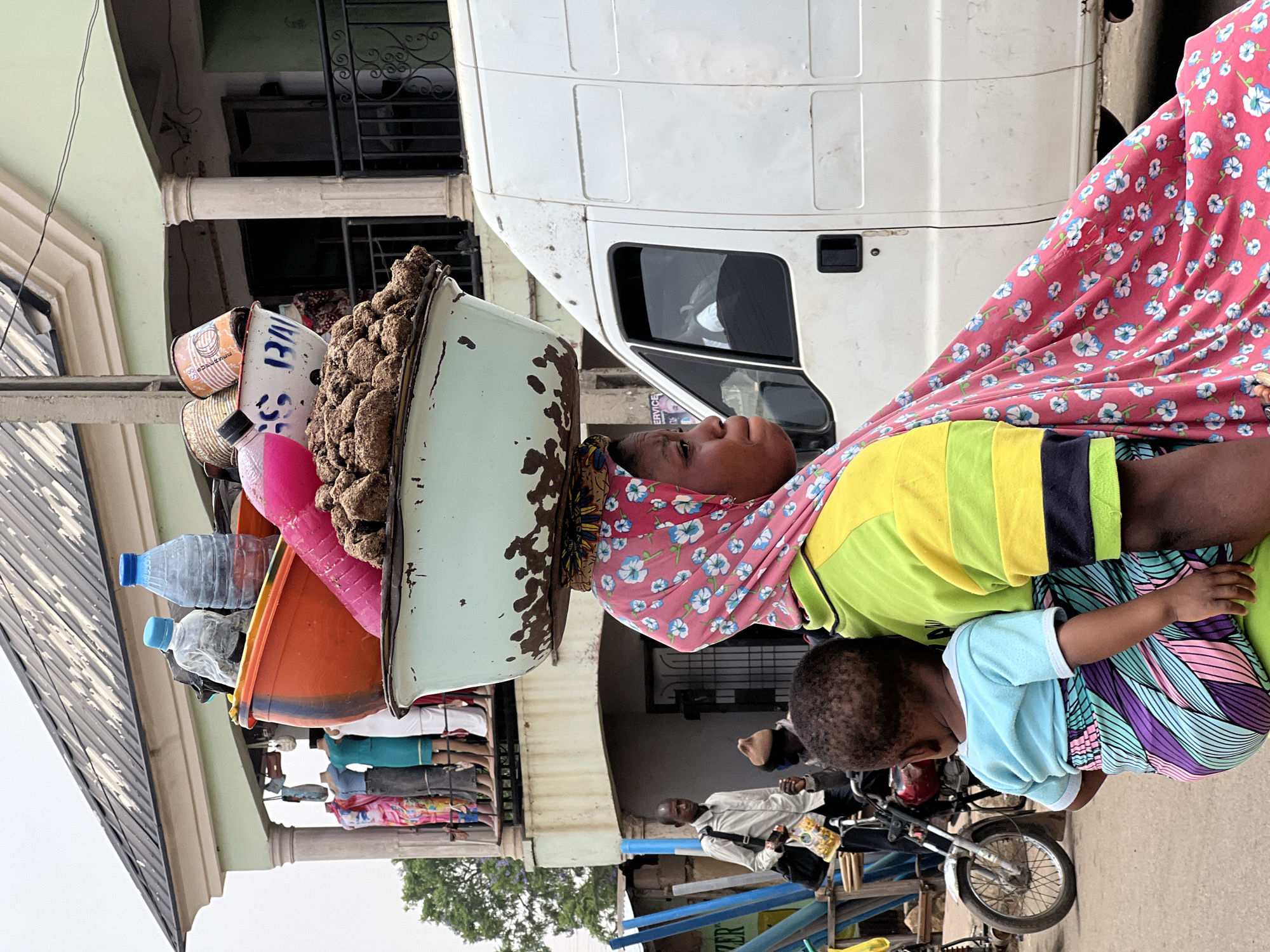 In Bukuru, Nigeria, a woman balances a large, heavily laden metal tray on her head. The tray contains a mix of goods, including a pile of brown, round food items, plastic containers, and bottles. She is wearing a floral patterned headscarf and a multi-colored top. A child clings to her side. A white van is parked beside her. In the background, there are other people and a motorcycle. Buildings and elements of urban infrastructure are visible, suggesting a busy street scene.