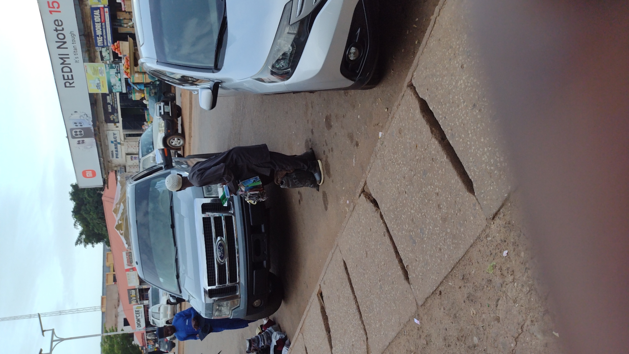 The image depicts a street scene in Jos, Nigeria. A man wearing a white cap and a dark robe stands beside a gray Ford F-150 pickup truck. He is holding a collection of items, possibly keychains and small accessories, in his right hand and appears to be interacting with the driver or passenger of the truck. Another person, dressed in a blue outfit and a cap, stands on the left side of the pickup truck, also near some bags of merchandise. In the background, buildings with signs are visible, including one that reads "REDMI Note 15" and another with "PHARMACY". Several other vehicles are present on the street, and the ground is paved with concrete slabs, some of which are cracked. The overall atmosphere suggests a busy commercial area.