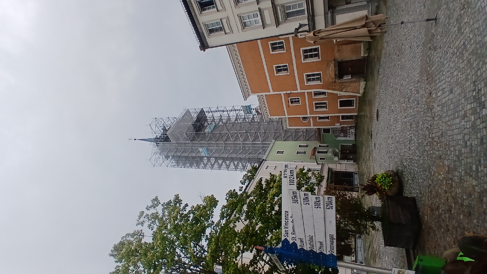 This is a portrait-oriented, full shot of a European town square in Pfarrkirchen, Germany, on an overcast day. The primary focus is a tall building undergoing extensive scaffolding work, likely a church or historic tower, with a spire visible at its peak. Surrounding it are several buildings with colorful facades – one orange and two green – featuring traditional European architecture with multiple windows. The ground is paved with cobblestones, and a large, closed patio umbrella stands near one of the buildings, suggesting an outdoor seating area for a cafe or restaurant.
In the foreground, a large, leafy tree partially obscures the view, and to its right, a series of blue directional signs are visible. These signs list several town names and their distances, indicating this is a public thoroughfare. The text on the signs is partially obscured but includes "San Vincenzo," "St. Rémy de P.," "Ruswil," "Buttisholz," "Ettiswil," and "Grosswangen," along with corresponding distances in kilometers.
The overall atmosphere is subdued due to the overcast weather. The scaffolding suggests ongoing construction or renovation, which is the main activity occurring in terms of the scene's visual narrative. No people are clearly visible in the image. The time of day appears to be daytime, but the lack of direct sunlight makes it difficult to pinpoint precisely. The location is identifiable by the town square setting and the German town name.