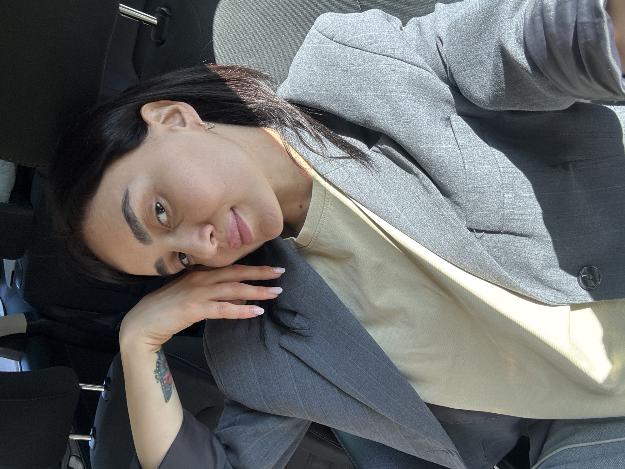 A brunette woman with fair skin and arched eyebrows is reclining in what appears to be a car seat. She is wearing a gray blazer over a beige top. Her left arm is bent with her hand near her face, and a butterfly tattoo is visible on her left forearm. The background suggests the interior of a car with headrests visible. The scene is likely not in the city of Tbilisi, Georgia, as there is no evidence in the picture to suggest that.