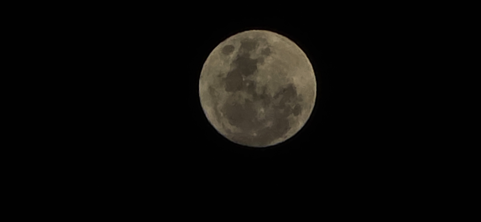 The image shows a full moon in a black sky. The moon's surface texture is visible, with varying shades of grey and darker patches. The location is unknown and cannot be ascertained by the visual alone.