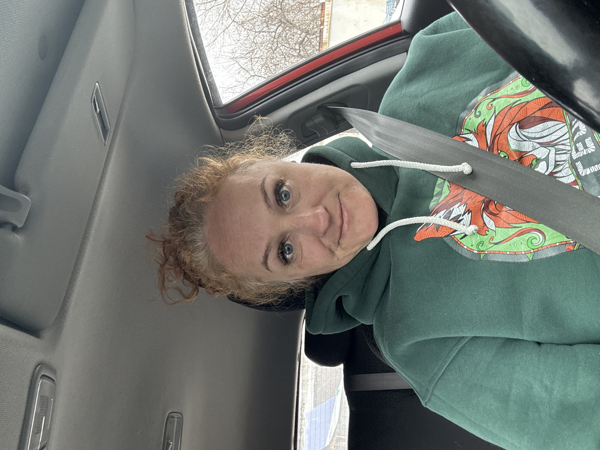 A woman with curly red hair is wearing a green hooded sweatshirt with a colorful design and a seatbelt. She is inside a red car. Through the car window, trees and a building are visible. Based on the request for location, it can be assumed that the scene is in the city of Chaikovsky, Russia.
