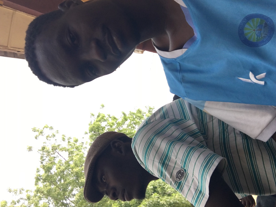 The image displays two dark-skinned males positioned outdoors in Pindiga, Nigeria. The individual on the right, facing forward and slightly left, looks directly at the camera. He wears a light blue and white short-sleeved shirt with a white "k" logo on the left chest and a circular emblem on the right chest. To his left, a second male is visible in profile, wearing a brown flat cap and a white polo shirt with thin teal horizontal stripes and a "DISCUS" patch on the left sleeve. He is looking upwards and to the left. The background consists of a bright, overcast sky, green tree foliage in the upper left, and faint overhead wires. A reddish-brown structure is partially visible along the top right edge of the frame. The scene is illuminated by bright, diffused natural light.
