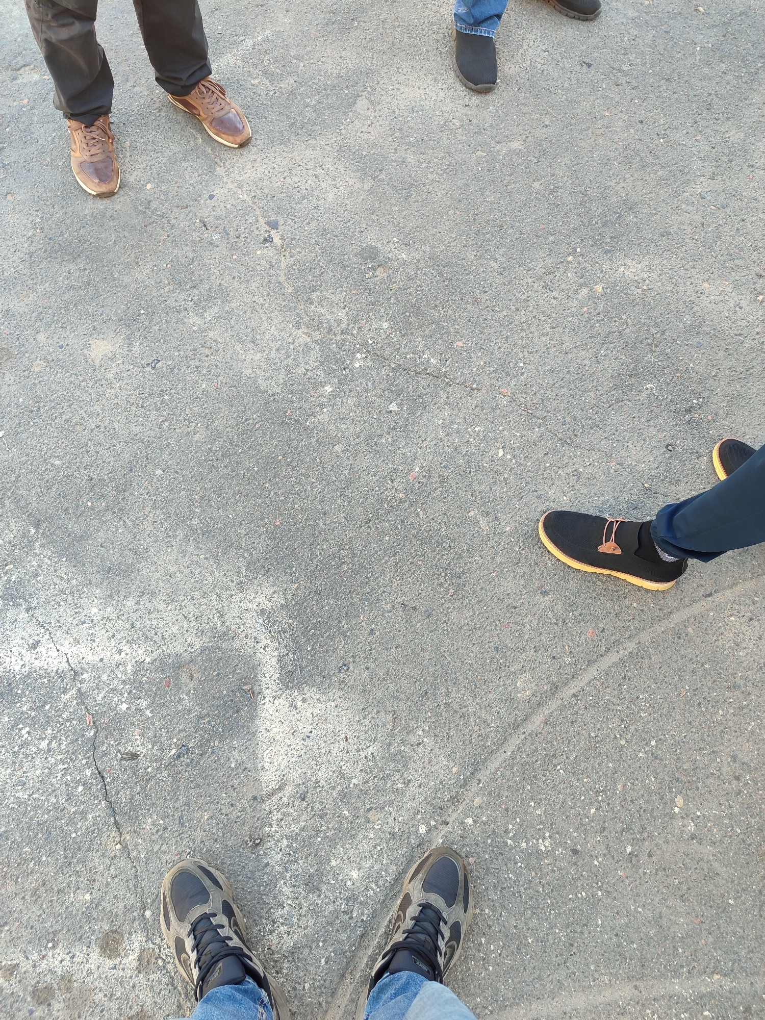 The image shows a ground-level view of a concrete surface, presumably a sidewalk or pavement, in an unknown urban location. Four individuals are visible, represented only by their lower legs and footwear. The individuals are spaced apart, creating a circular formation. The clothing consists of denim jeans and dark pants. The footwear varies: one pair of dark gray shoes, one pair of brown leather shoes, one pair of black shoes with tan soles and a brown accent, and one pair of grey athletic shoes. The concrete surface is marked with cracks and variations in texture. The lighting suggests daytime. No other objects or people are in view. There are no visible buildings, landmarks, or identifying information of any kind.