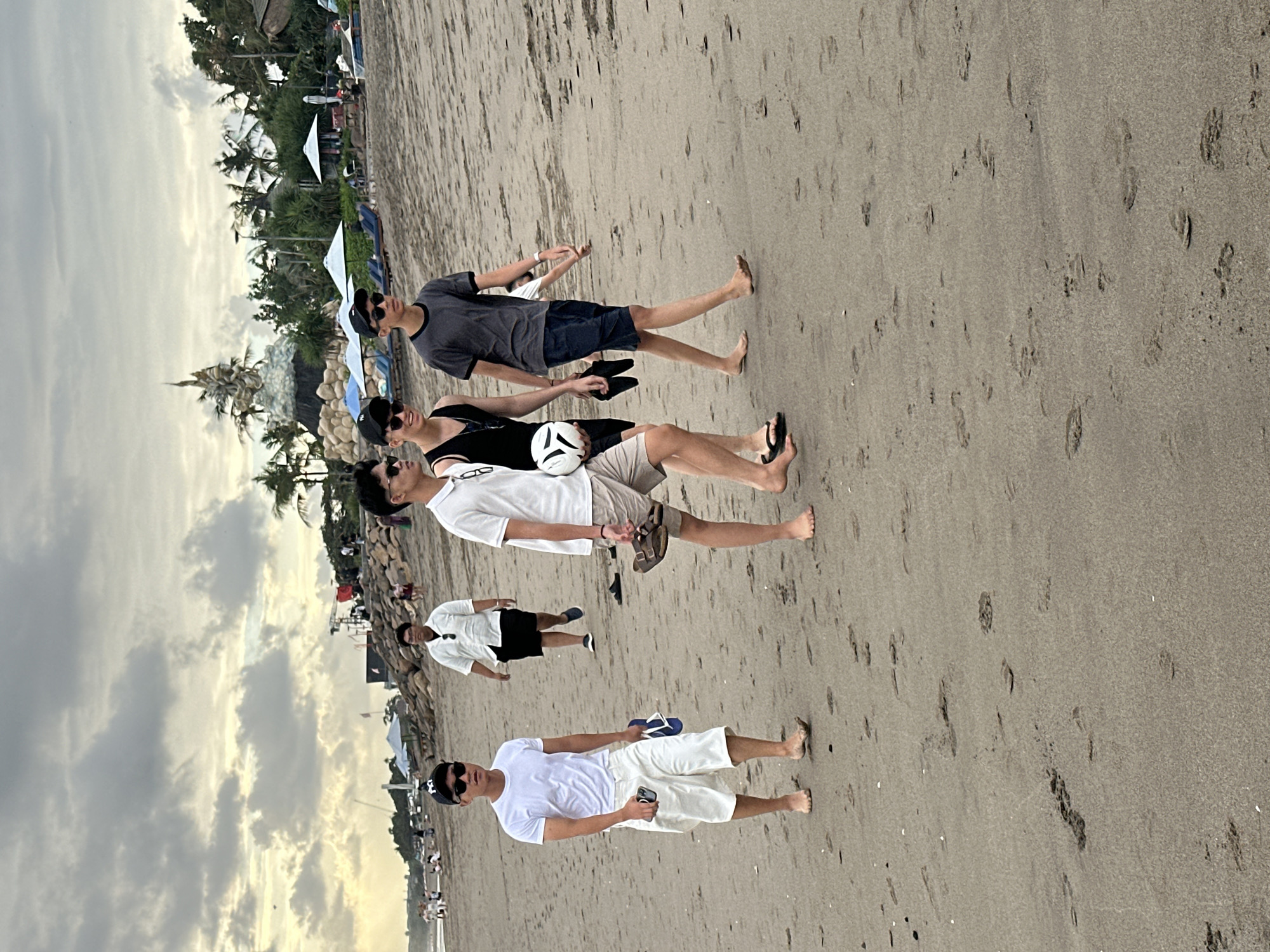 The image depicts a group of four young men walking barefoot on a sandy beach in North Kuta, Indonesia, under a cloudy sky. The man in the foreground on the left wears a white t-shirt and white shorts, carrying a phone and blue flip-flops in his hands. To his right, a man in a black tank top and shorts carries a white ball and black flip-flops. Next to him, another man wears a grey t-shirt and blue shorts. A fourth man, in a white polo shirt and black shorts, is visible further in the background, walking in the opposite direction. The beach features footprints in the sand, and in the distance, there are palm trees, umbrellas, lounge chairs, and a large decorative structure. The overall atmosphere appears casual and relaxed.