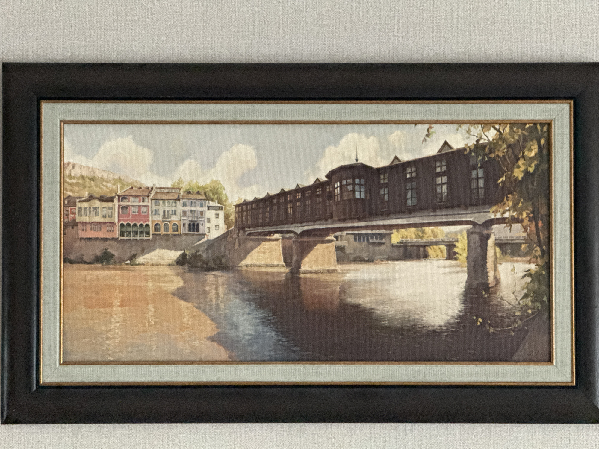 A framed painting depicts a scene of the Covered Bridge in Lovech, Bulgaria. The bridge, a wooden structure with enclosed shops or rooms along its length, spans a river reflecting the bridge and surrounding buildings. On the left bank are buildings with arched windows and varying colors, with a mountain in the background. The river flows in the foreground, with the right side having foliage. The painting is framed in a dark matte frame with a lighter inner frame.