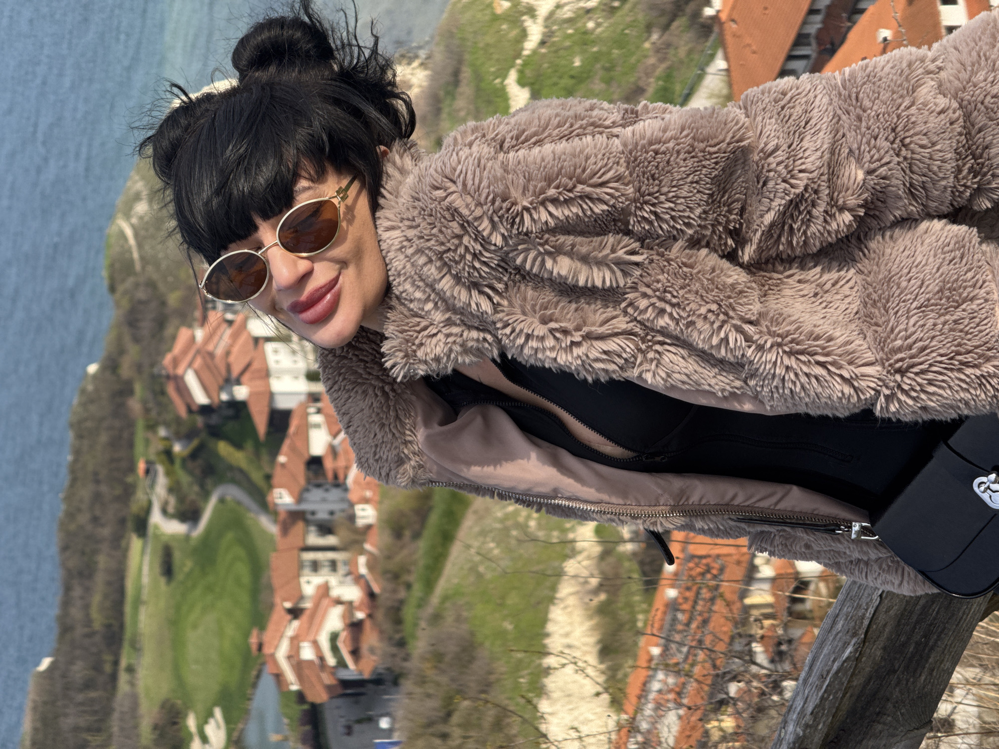 A woman wearing sunglasses and a furry coat is standing outdoors on a wooden structure. Behind her is a view of a coastal area with buildings and greenery. The scene suggests an overlook or elevated viewpoint. The city of Topola in Bulgaria is not identifiable from the image.