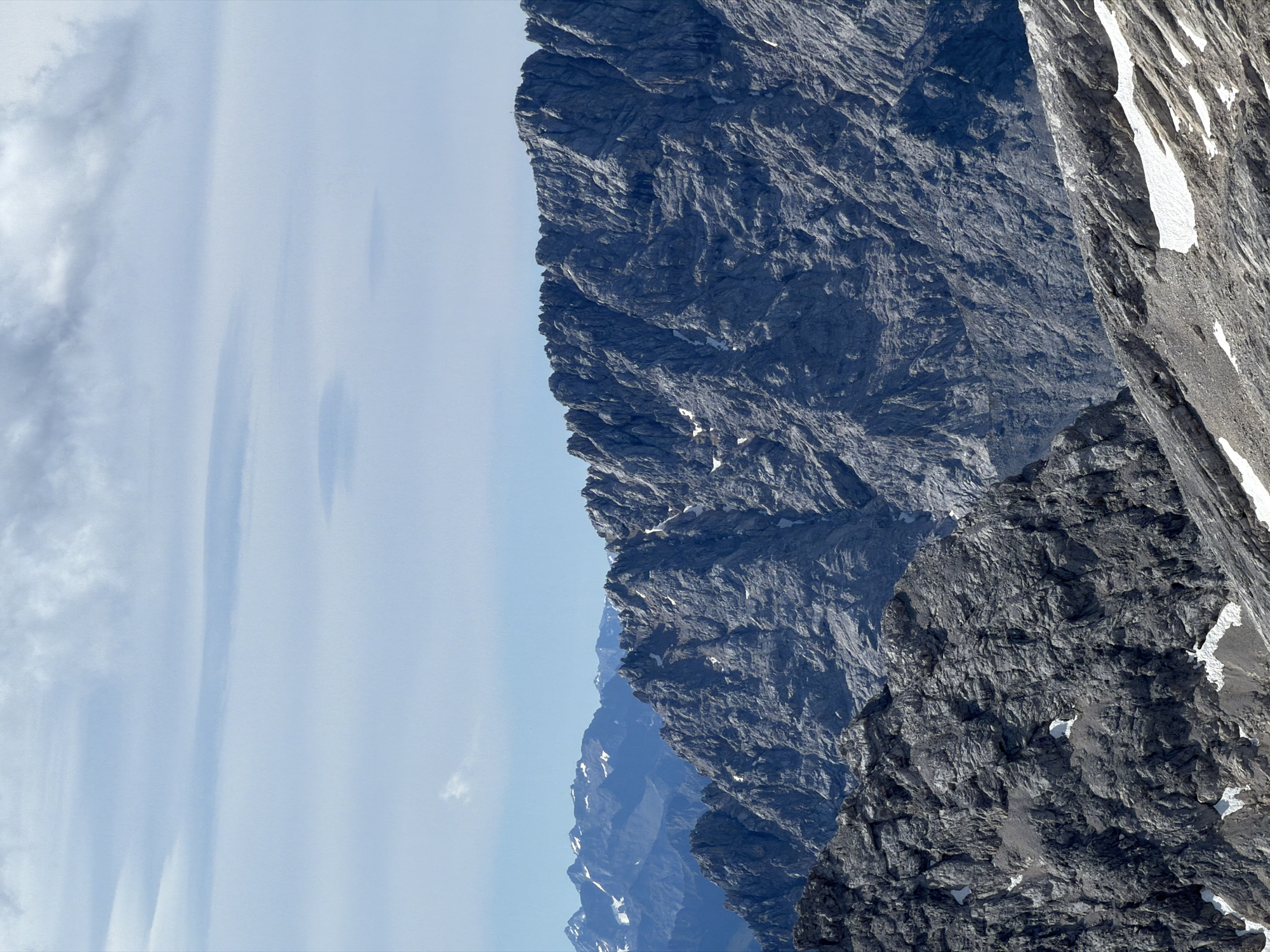 The image depicts a mountainous landscape, characteristic of the Bavarian Alps near Garmisch-Partenkirchen, Germany. The foreground and mid-ground are dominated by jagged, rocky mountain ranges, with visible textures and striations indicating geological formations. Patches of snow are scattered across the slopes, particularly in the lower parts of the frame. In the background, a series of progressively receding mountain ranges fade into a hazy blue, with the furthest peaks showing hints of snow cover. The sky above is a pale blue, partially obscured by scattered, wispy clouds. The scene is a natural environment, devoid of any human presence or man-made structures.