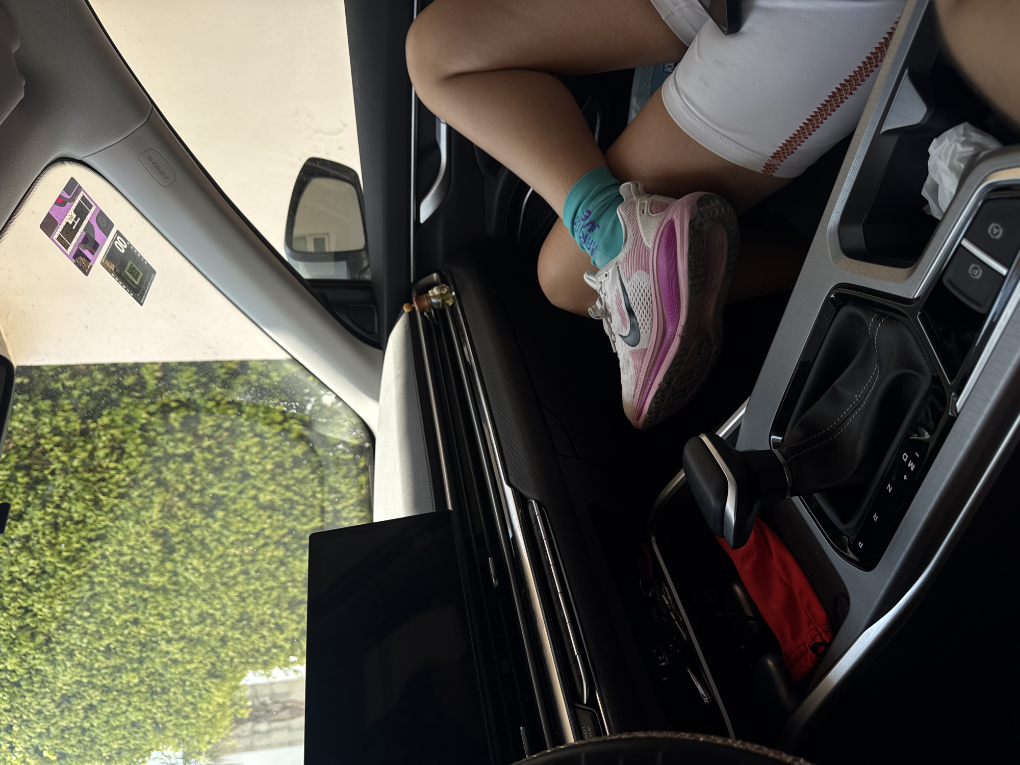 The image shows the interior of a car, with a person's legs visible. The person is wearing white shorts, a light blue sock, and a pink and white running shoe. The gear shift and console of the car are prominent, and a red object is in the storage area below the shift. The car is parked, and trees are visible through the car window. There are no clues in the image to specifically determine the location is Heroica Puebla de Zaragoza, Mexico.