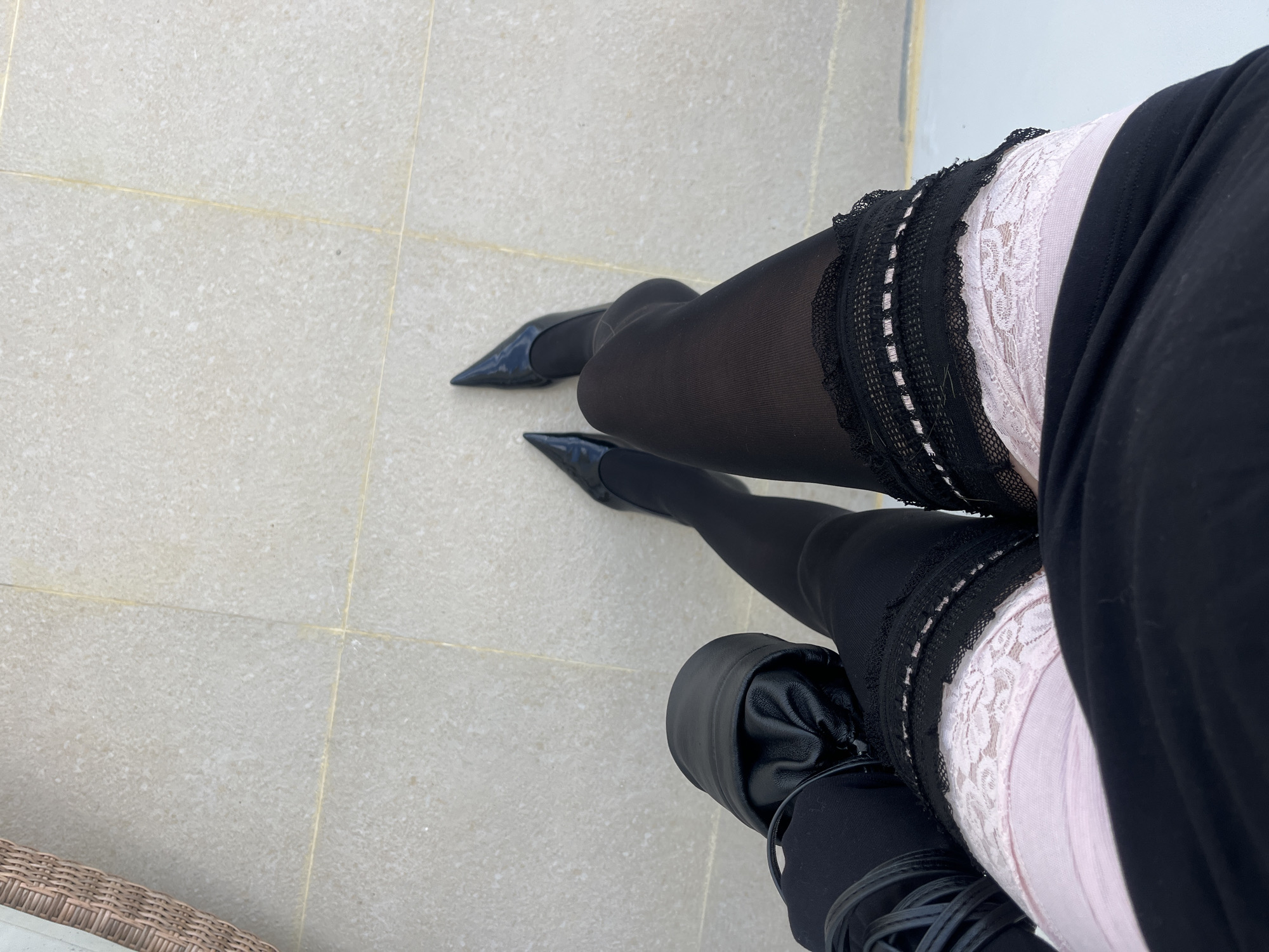 A high-angle shot captures a person's lower body, focusing on their legs and footwear, set against a light-colored tiled floor. The individual is wearing sheer black tights or stockings, which transition into pointed black patent leather high heels on both feet. Peeking out from beneath a black garment, possibly a skirt or dress, are light pink, lace-topped thigh-high stockings or garters, layered beneath the sheer black hosiery. The intricate lace pattern on the pink undergarment is distinctly visible, contrasting with the solid black attire above it.
On the right leg, a black, textured, possibly leather-like covering, which appears to be part of a boot or a leg accessory, is visible near the ankle and lower calf. On the left side of the frame, a portion of a woven, wicker-like furniture piece, likely a chair, can be seen in the bottom corner.
The floor is made of light beige or off-white tiles with visible, slightly darker grout lines, suggesting an outdoor patio or balcony setting. The bright, even lighting indicates it is daytime. No faces or upper bodies are visible, and there is no discernible text in the image. The overall impression is one of stylish and coordinated attire, presented from a unique overhead perspective in Kuta Selatan, Indonesia.