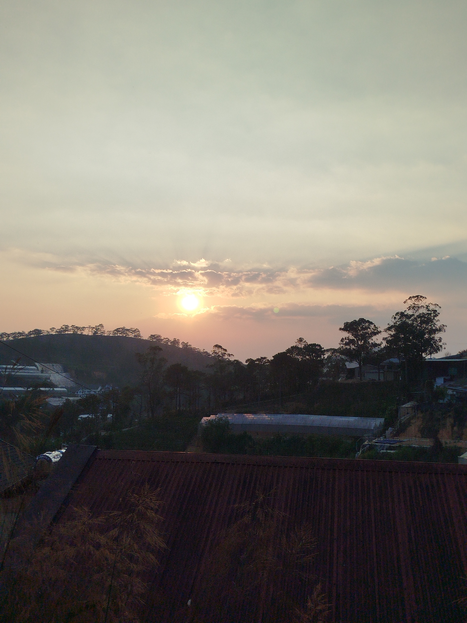 The image depicts a sunset over a hilly landscape in Lạc Dương, Vietnam. The sun is partially obscured by clouds, casting a warm glow across the sky. The foreground is dominated by a corrugated metal roof. Trees dot the landscape, with some appearing as silhouettes against the setting sun. Structures, possibly greenhouses, are also visible. The scene appears tranquil and still.