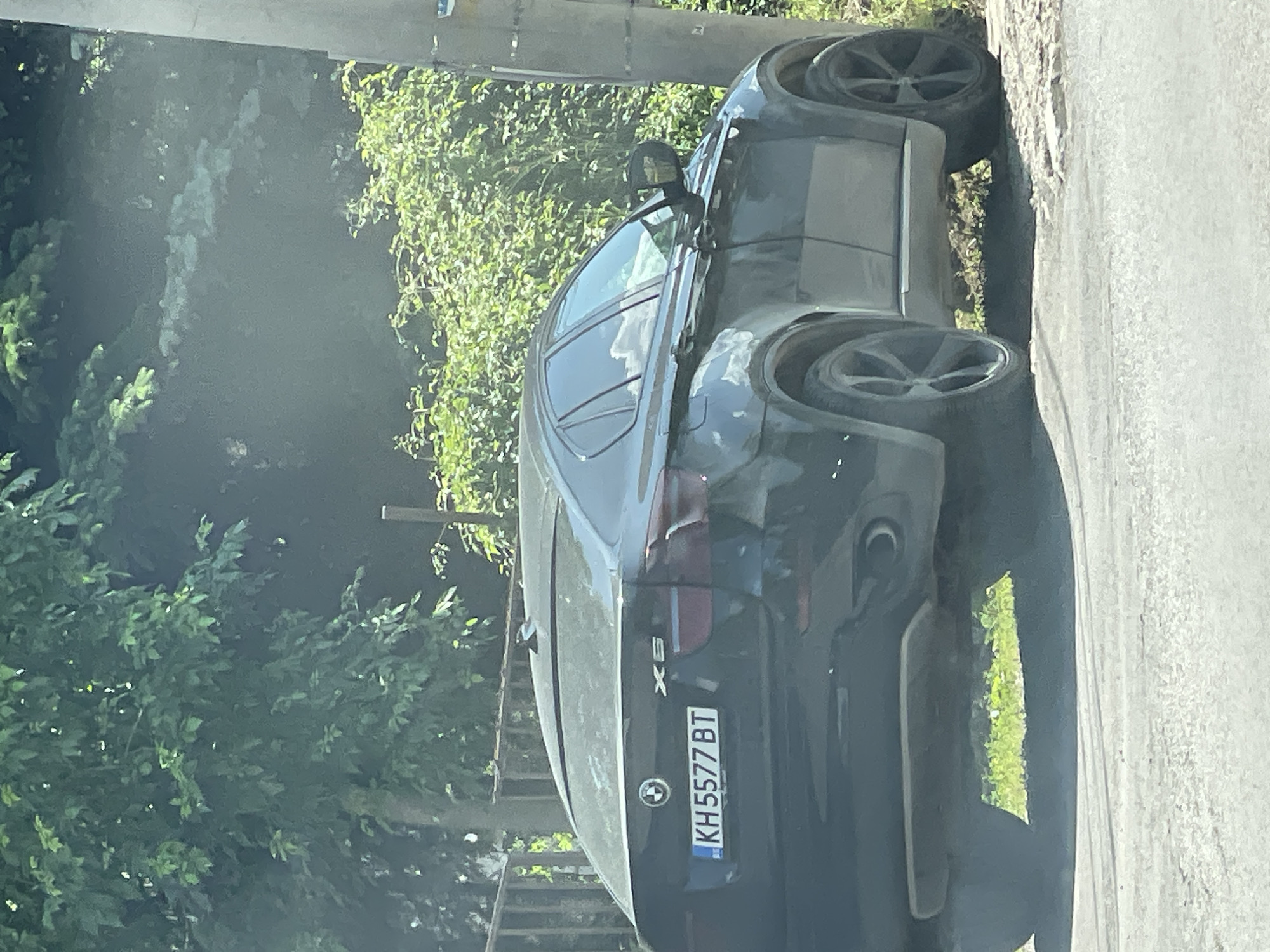 The image captures the rear-left side of a black BMW X6 parked on a paved surface next to a grassy embankment. The car's license plate reads "KH5577BT". The background features dense green foliage, with a section of a wooden fence visible to the left and a utility pole on the right. The lighting suggests it is daytime, with sunlight casting shadows and creating reflections on the car's surface. No people are visible in the frame.