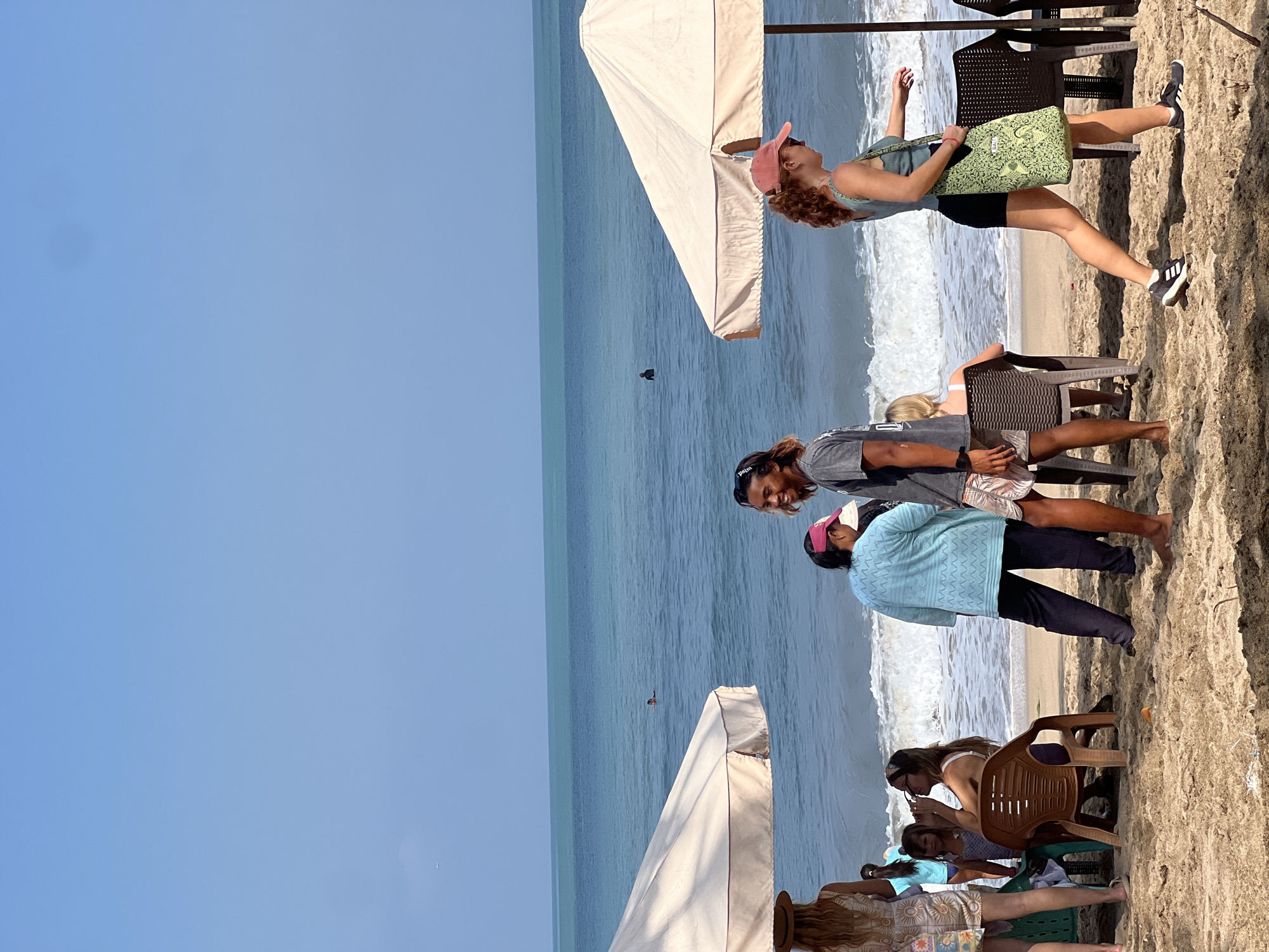 A group of people are on a sandy beach in Kuta, Indonesia, with the ocean and sky in the background. The sky is clear and blue, with a thin line of turquoise water meeting the lighter blue ocean. Waves are breaking on the shore.
On the left side of the image, a woman with blonde hair wearing a patterned dress and a hat stands near a beige umbrella. A young girl with blonde hair is seated on a green chair to her left. Further to the left, partially visible, are more people seated and standing.
In the center of the image, a man with long dark hair and a black t-shirt is smiling and looking towards the right. He is wearing patterned shorts. Next to him, a person in a blue top and dark pants is standing and looking down.
On the right side of the image, a woman with curly red hair, wearing a pink baseball cap, sunglasses, a teal tank top, and black shorts, walks towards the right carrying a green patterned tote bag. She is wearing athletic shoes. Behind her, another beige umbrella is visible, and chairs are placed around it. A woman with blonde hair is seated on one of these chairs, facing away from the camera.
The sand is light brown and appears dry, with some scattered debris. The overall scene depicts a relaxed atmosphere on a sunny day at the beach.