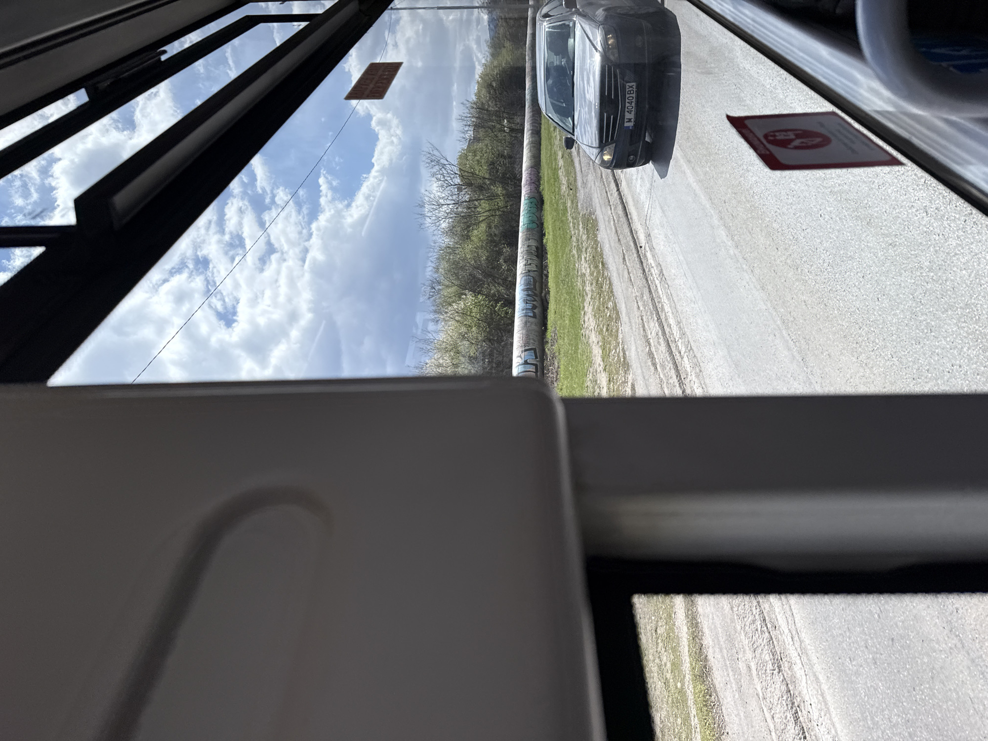 A view from the inside of a moving tram in Sofia, Bulgaria, shows a street with a parked car, a pole covered with graffiti, trees, and a cloudy sky. The car's license plate number is M 4040 BX. Parts of the tram's interior are visible.