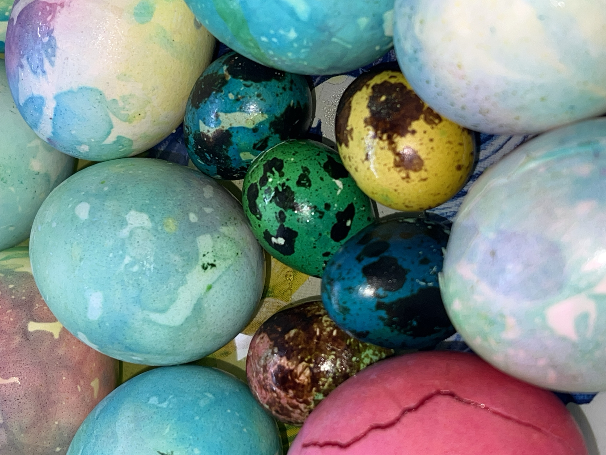 The image shows a close-up view of a pile of Easter eggs. The eggs are dyed in various colors including shades of blue, green, yellow, pink, purple, and white. Many have speckled or marbled patterns. One pink egg has a crack. The eggs are touching each other, indicating they are tightly packed. The image does not contain information to determine a location in the city of Noviy, Russia.