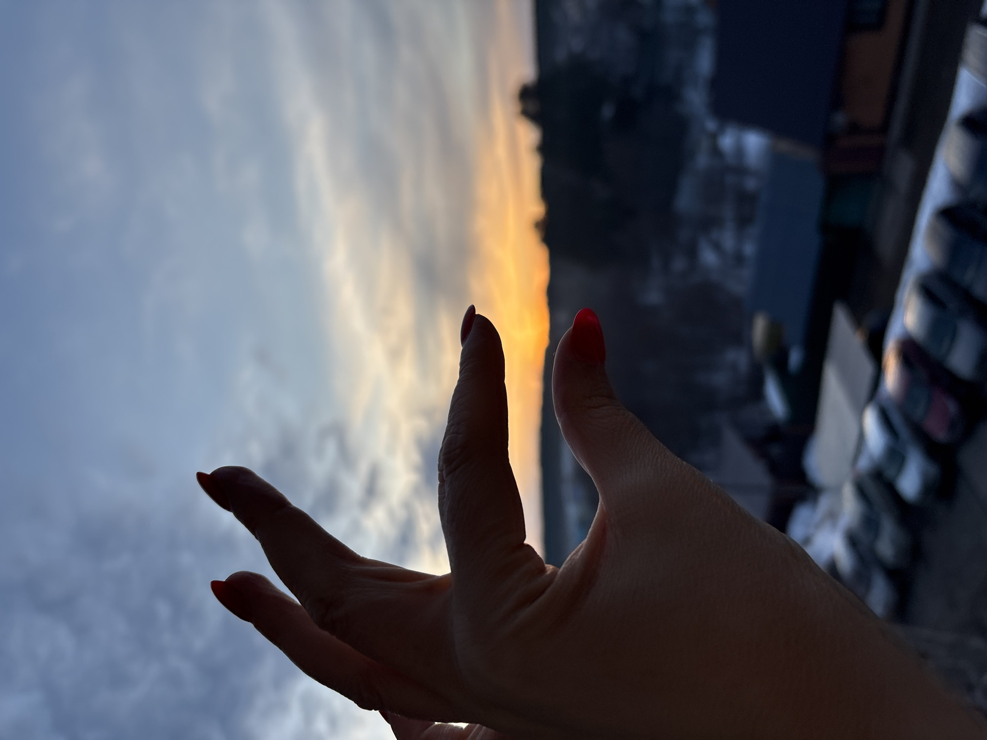 This image depicts a human hand silhouetted against a sunset or sunrise in Chaikovsky, Russia. The hand, with visible red-painted fingernails, is positioned in the foreground with the thumb and index finger extended, appearing to "frame" or "pinch" the sun. The sky transitions from bright orange and yellow at the horizon, where the sun is visible, to cooler blue and gray tones with scattered clouds overhead. In the mid-ground, a row of parked cars, partially covered in what appears to be snow, is visible. Behind the vehicles, several dark-roofed buildings are discernible, followed by a silhouette of distant trees and possibly hills along the horizon. The ground in the mid-ground shows evidence of snow, indicating a cold weather environment.