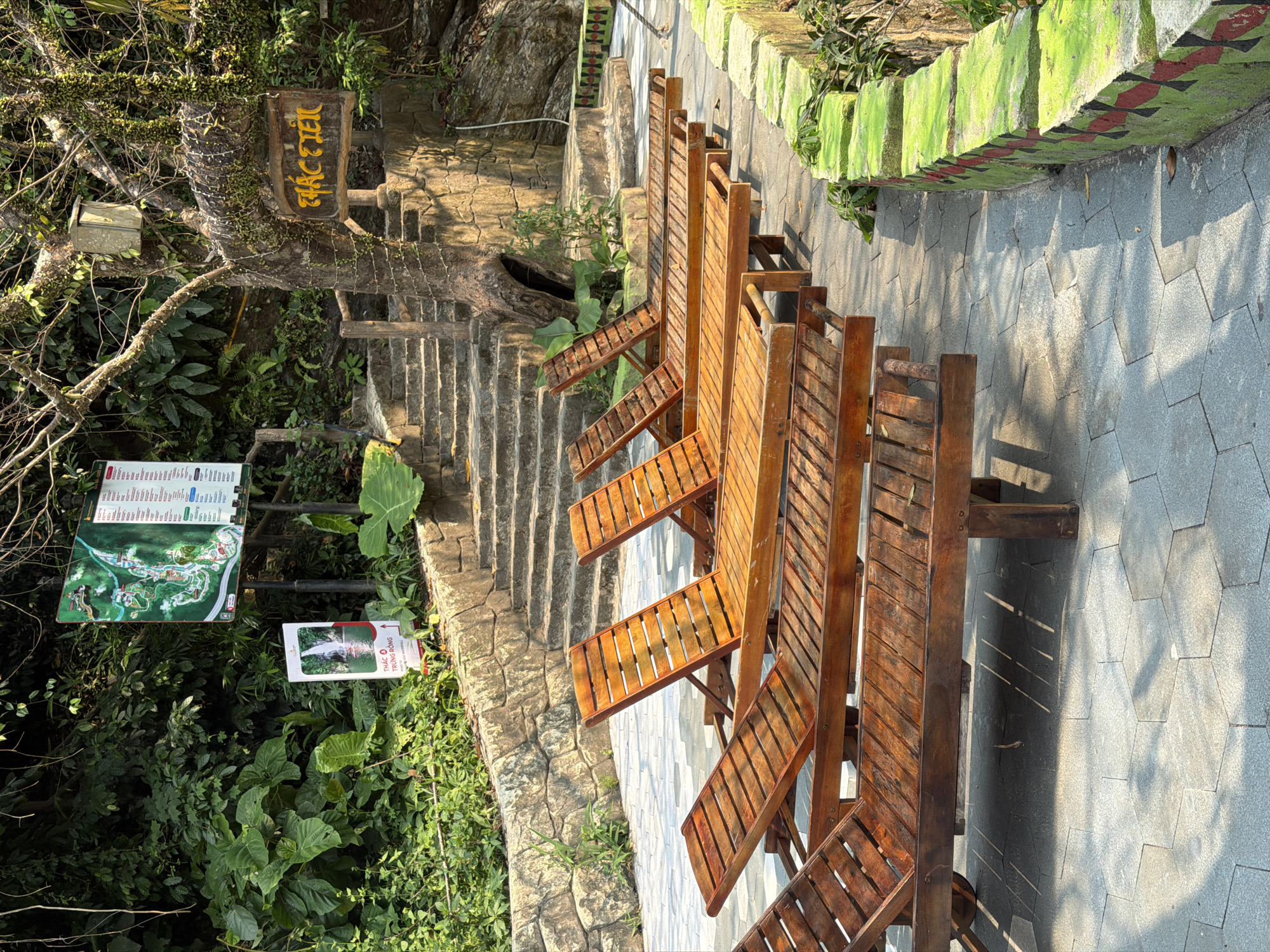 The image depicts a seating area at the Dragon Bridge Waterfall (Thác Rồng) in the Bà Nà Hills resort near Đà Nẵng, Vietnam. Several wooden lounge chairs are arranged on a paved walkway. To the left, stone steps lead up to the waterfall and surrounding area. Signage and lush vegetation are visible.