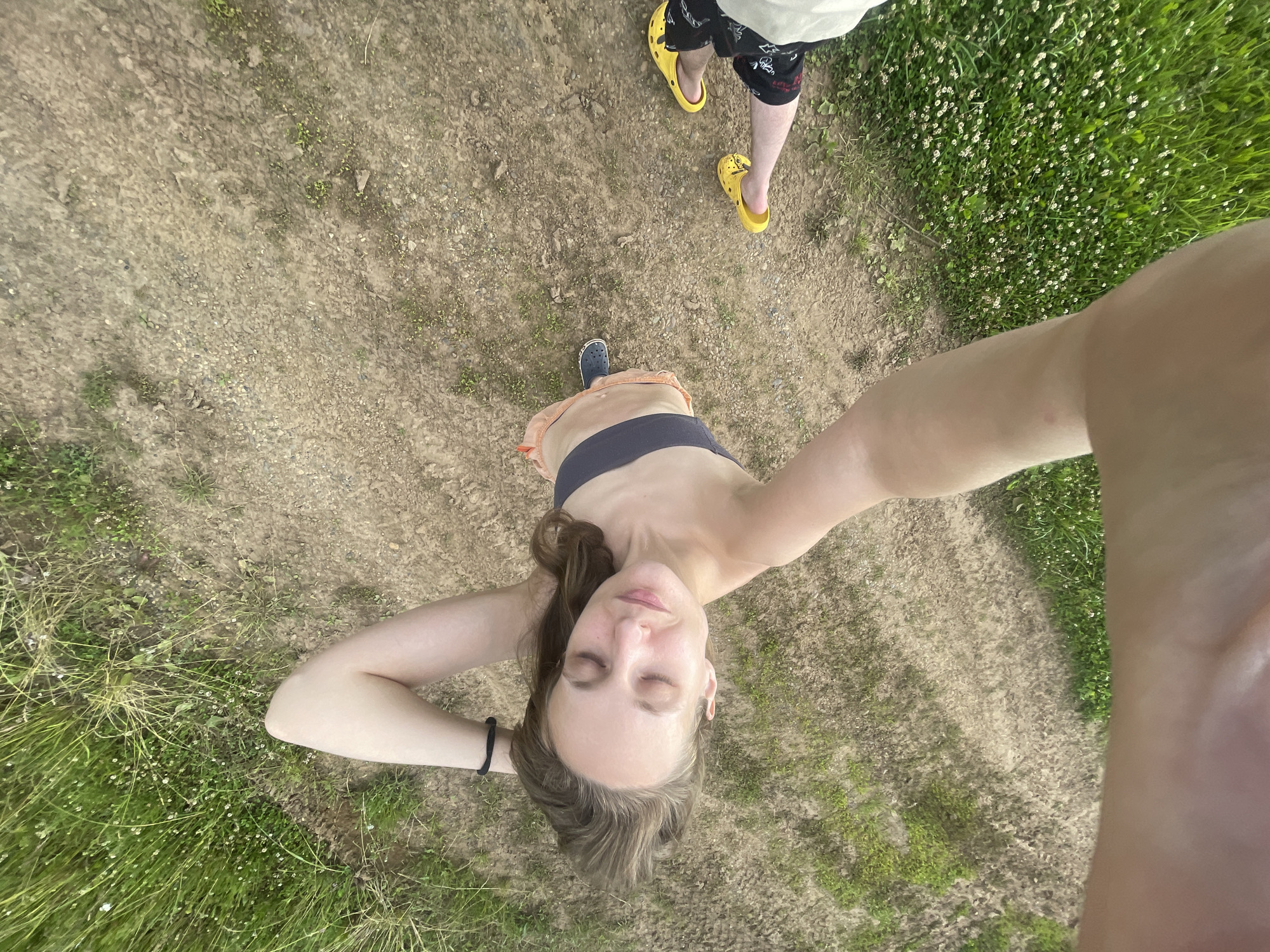 This is a downward-facing, wide-angle selfie taken outdoors. The primary subject is a young woman with light brown hair, wearing a dark sports bra and orange shorts. She is lying on her back with her head tilted back, eyes closed, and her arms behind her head. A black hair tie is visible on her left wrist.
In the background, the ground appears to be a dirt path with scattered small stones and patches of green grass and clover. To the right, a dense patch of green grass with small white flowers is visible.
Further up in the frame, the legs of another person are visible, wearing black shorts with a pattern and bright yellow Crocs. This person is standing on the dirt path.
The lighting suggests it is daytime, possibly late afternoon given the warm tones. The overall impression is one of relaxation and being outdoors in a natural setting, possibly a park or a trail in Kuyeda, Russia. No text is visible in the image.