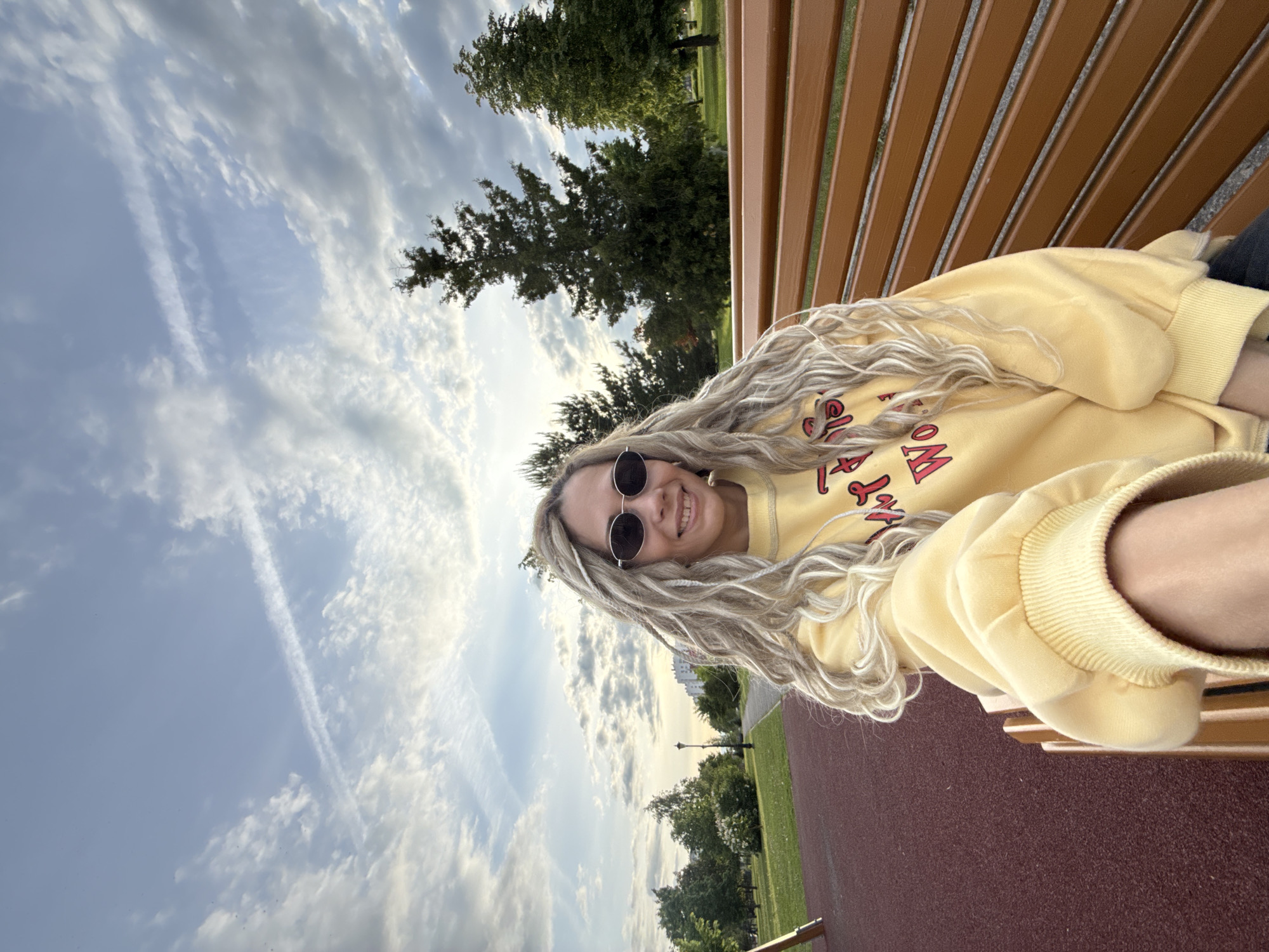 The image displays a young woman posing for a photo, taken from a slightly tilted perspective, as she sits on a wooden slatted park bench. She is smiling broadly at the camera, wearing dark, round sunglasses and a bright yellow long-sleeved sweatshirt with red, stylized text partially visible on the front (e.g., "OM" or "BOM"). Her long, light-blonde hair is styled in numerous dreadlocks or braids, flowing over her shoulders and the bench.
The setting is an outdoor park or green space, characteristic of Заволжье, Russia. The background features a partly cloudy sky with patches of blue, and a prominent, straight airplane contrail stretching across the upper left. Various green trees, both tall and dense, are scattered throughout the park, providing a natural backdrop. A reddish-brown pathway or recreational surface is visible in the foreground and midground. In the distance, a lamp post and faint outlines of buildings can be seen, indicating an urban park environment. The lighting suggests it is daytime, possibly late afternoon or early evening, with soft, natural light. The woman's cheerful expression conveys happiness and relaxation in the pleasant outdoor setting.