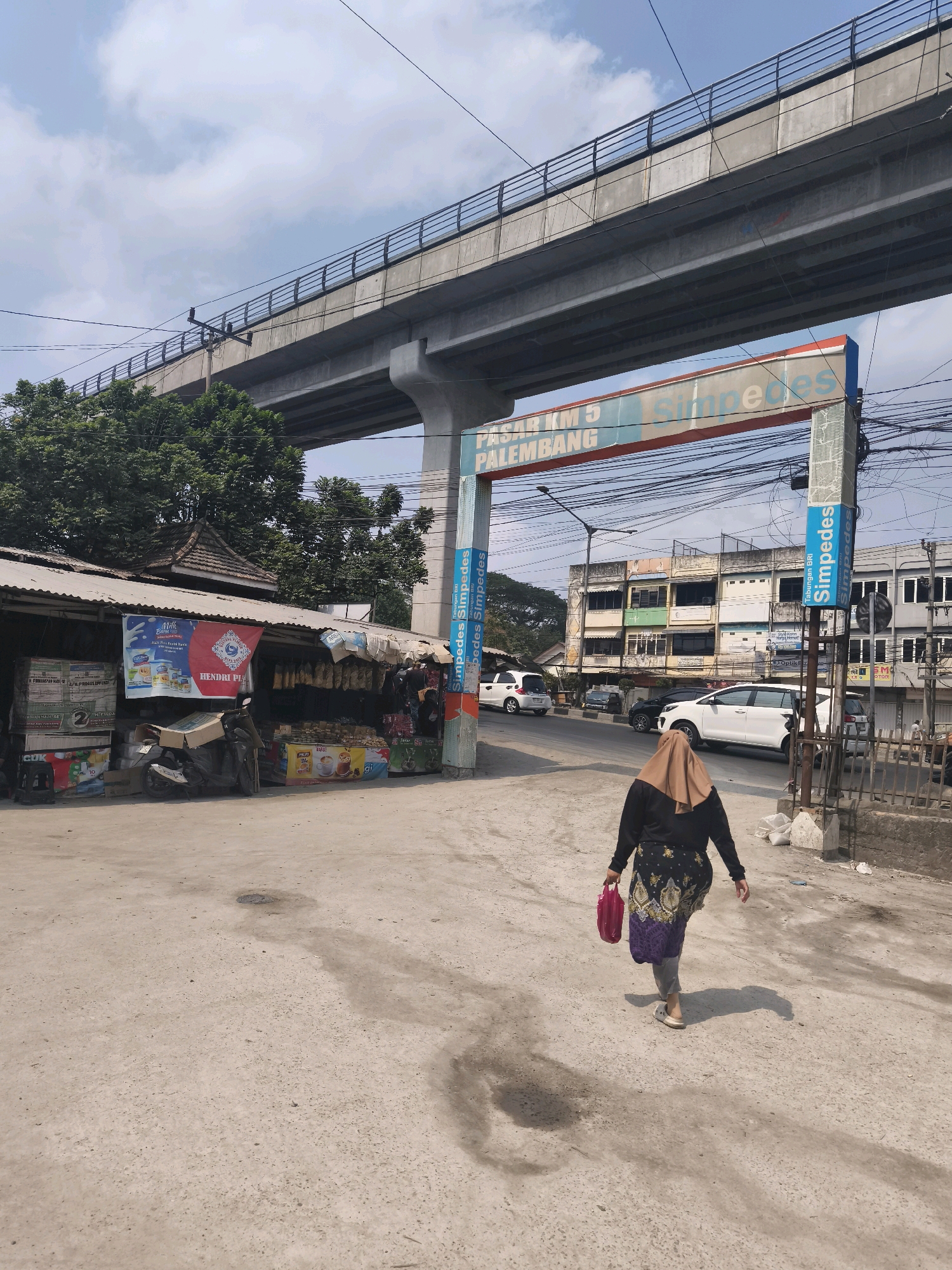 The image shows a street scene in Palembang, Indonesia, under an elevated highway. A woman wearing a hijab and a black top with a patterned skirt is walking away from the camera on a dirt and asphalt road. She is carrying a pink plastic bag. To the left, there is a roadside market stall with a corrugated metal roof, displaying various goods including bottled drinks and snacks. A motorcycle is parked in front of the stall. In the background, several buildings line the street, and a white car and other vehicles are visible on the road. An archway with the text "PASAR KM 5 PALEMBANG" and "Simpedes" is present, indicating the location as Market Kilometer 5 in Palembang. The sky is partly cloudy.