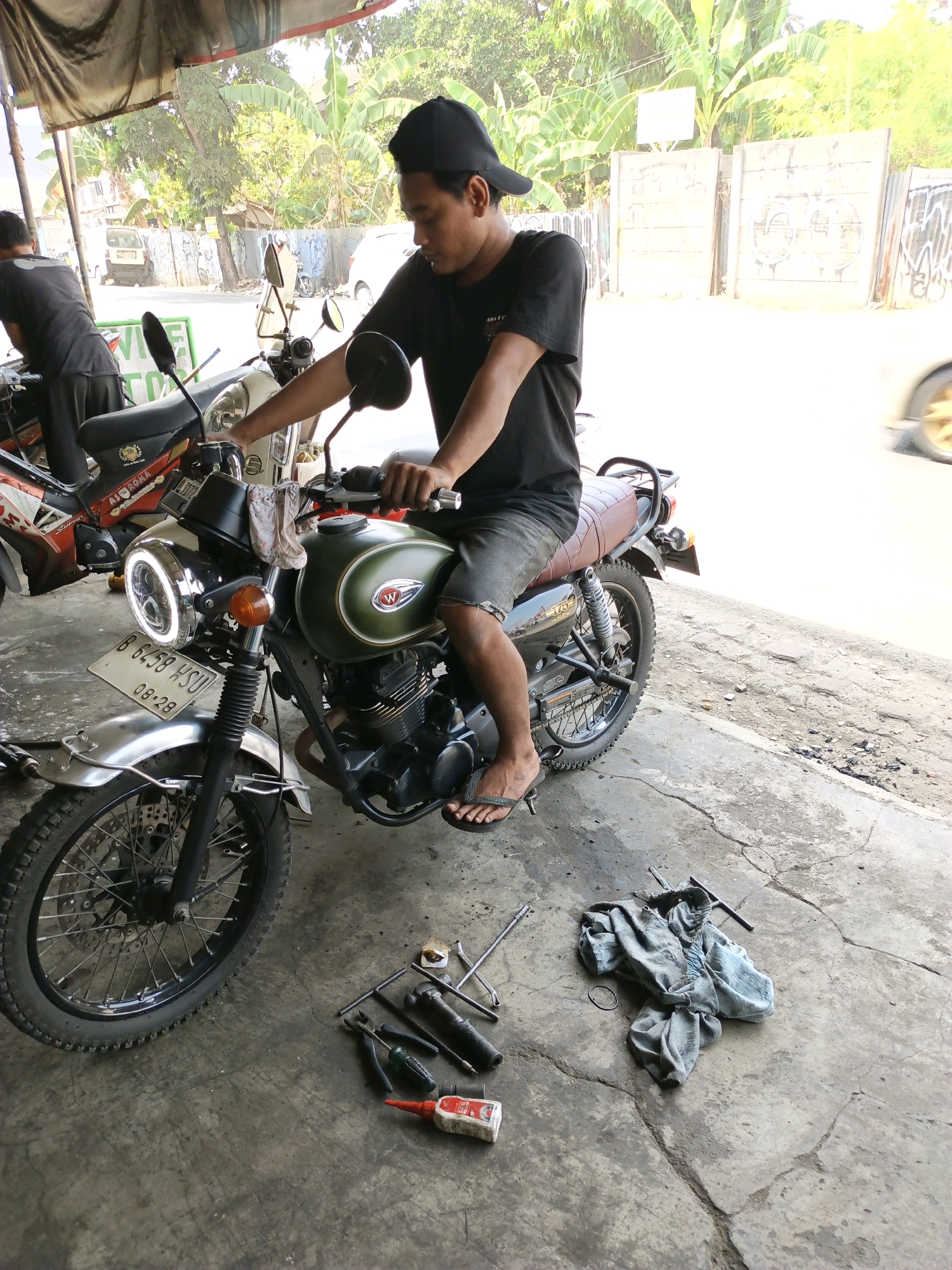 An Indonesian man wearing a black t-shirt, black shorts, and a black baseball cap is sitting on a green motorcycle with a brown seat. He is positioned in the driver's seat, with his right hand on the handlebar and his left hand resting near the controls. A stained cloth is draped over the handlebar. The motorcycle has a white license plate that reads "B 6458 HSU 08-28". In front of the motorcycle, on the concrete floor, are various tools including pliers, screwdrivers, a small bottle of red liquid, and a pile of blue rags. To the left of the main motorcycle is another motorcycle, partially visible. The scene appears to be an outdoor repair shop or garage. In the background, there is a street with trees and buildings. The man is looking down at the motorcycle. The overall lighting suggests it is daytime. The location is identified as South Tangerang, Indonesia.