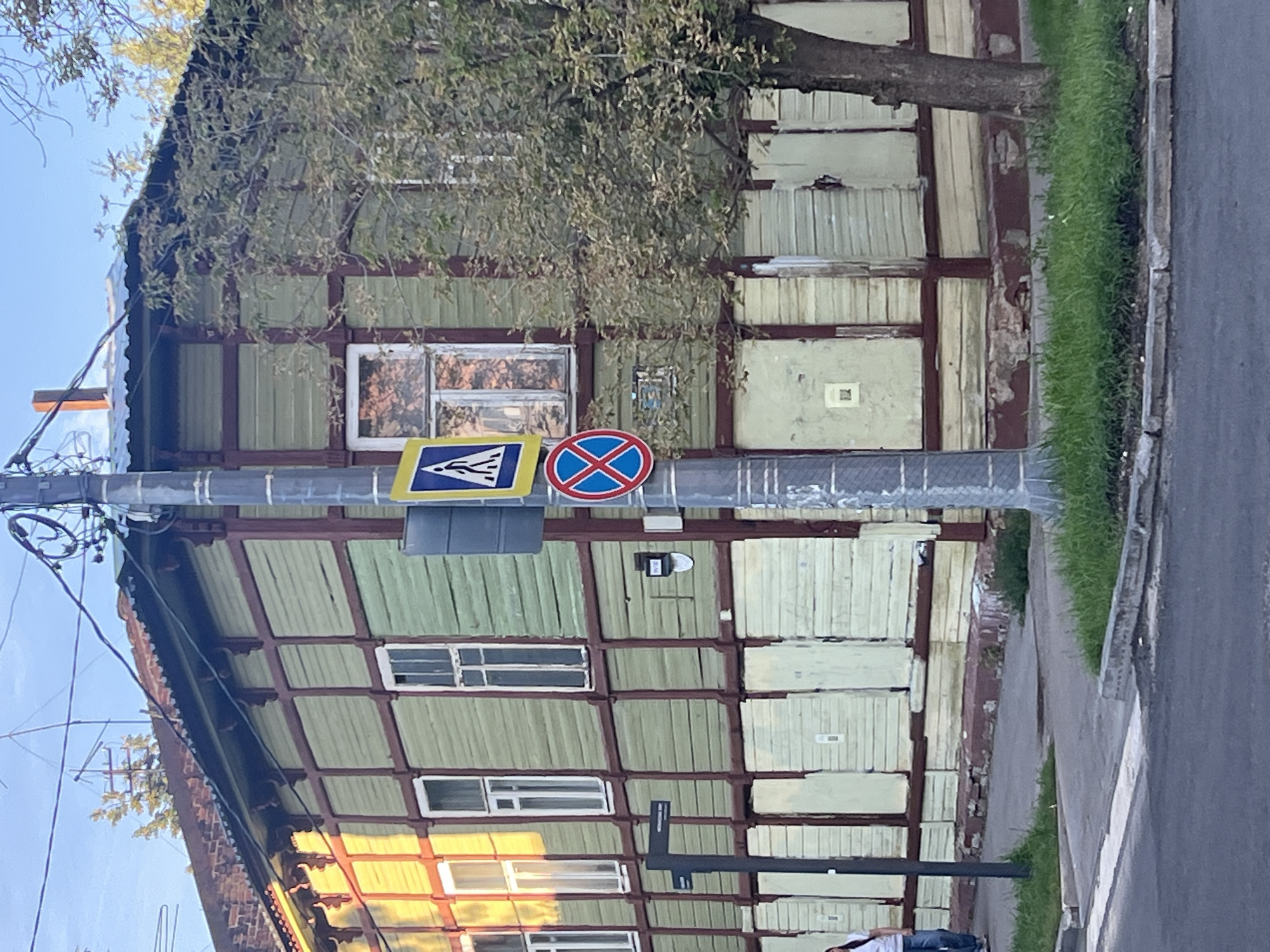 The image depicts a street scene in Nizhny Novgorod, Russia, focusing on a two-story wooden building with green siding and dark red trim. The lower story of the building is covered with light-colored wooden planks, suggesting it may be boarded up or have shutters. Several windows are visible on both stories, some reflecting the warm light of the setting sun.
A utility pole stands in the foreground, adorned with traffic signs. One sign is a blue square with a yellow border and a white pedestrian symbol, indicating a pedestrian crossing. Below it is a circular red sign with a blue diagonal stripe and a horizontal bar, signifying a prohibition, likely related to parking or stopping. A smaller black sign with white text is also attached to the pole, its text illegible.
To the left of the utility pole, a dark signpost with white directional arrows is positioned on the sidewalk. A person, partially visible from the waist down, walks on the sidewalk in the background, wearing jeans and a white t-shirt.
The building shows signs of wear and tear, with peeling paint and exposed wood on its foundation. Green grass grows along the edge of the sidewalk and the building's base. The street in the foreground is paved, with a visible crosswalk. The sky is a clear, pale blue, indicating either early morning or late afternoon light. The overall impression is a somewhat aged urban environment in Russia.