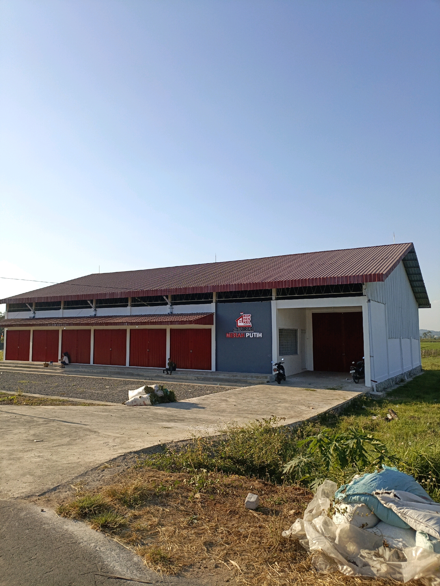 A long, rectangular, single-story building with a dark red, corrugated metal roof stands under a clear blue sky. The building has a light gray metal exterior with a white trim. The front of the building features a row of six red metal doors, each with a vertical white dividing line, set against a white facade. To the right of these doors is a recessed entryway with a large, open doorway revealing dark red double doors within. A small, dark gray rectangular sign with white text and a red logo is mounted on the left side of the building's facade. Two motorcycles are parked in front of the building. In front of the red doors, a gravel-covered area is visible. Further in the foreground, a concrete pathway slopes down to a grassy area with some dried brush and a pile of white and blue sacks. Two people are seated on steps in front of the red doors, and one person is seated on the ground to the right. The overall setting appears to be an industrial or storage facility in Rowosari, Indonesia, with a rural or semi-rural environment surrounding it.