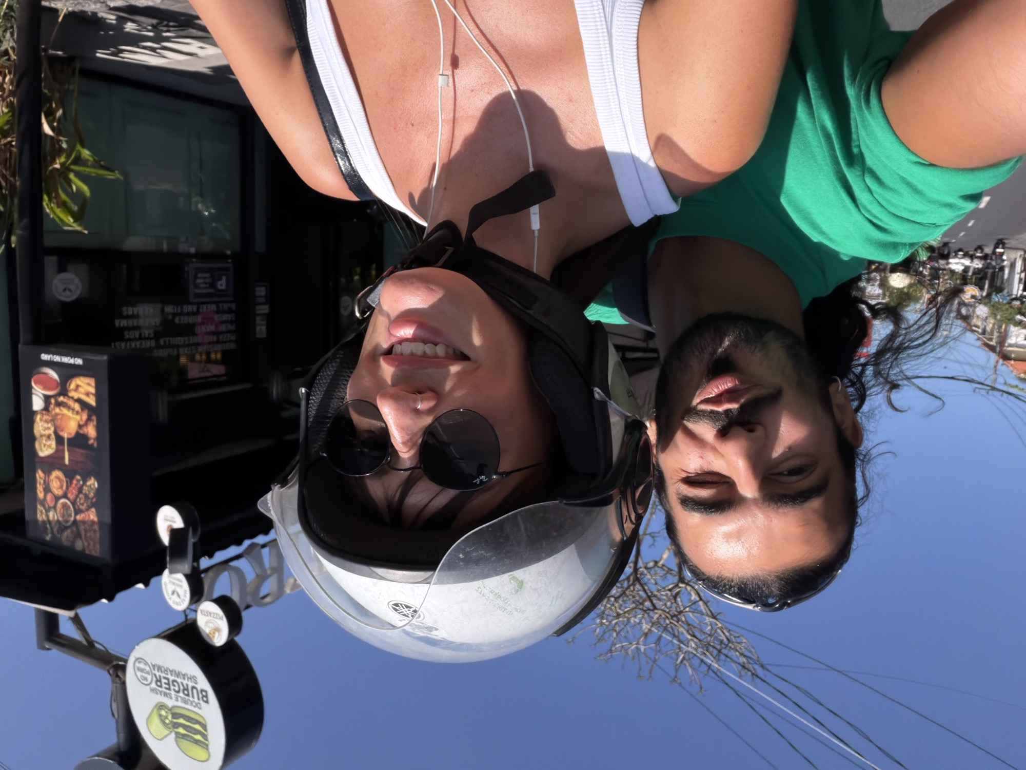 In the foreground, a man with dark, wavy hair and a beard is looking into the camera while riding on a motorbike. He is wearing a green t-shirt. To his right, a woman with dark hair is also looking at the camera, wearing a white tank top and a helmet with a clear visor. She is wearing sunglasses and has earbuds in. In the background, a sign for "Double Smash Burger Shawarma" is visible. The scene appears to be in a city with a clear blue sky. The location is identified as North Kuta, Indonesia.