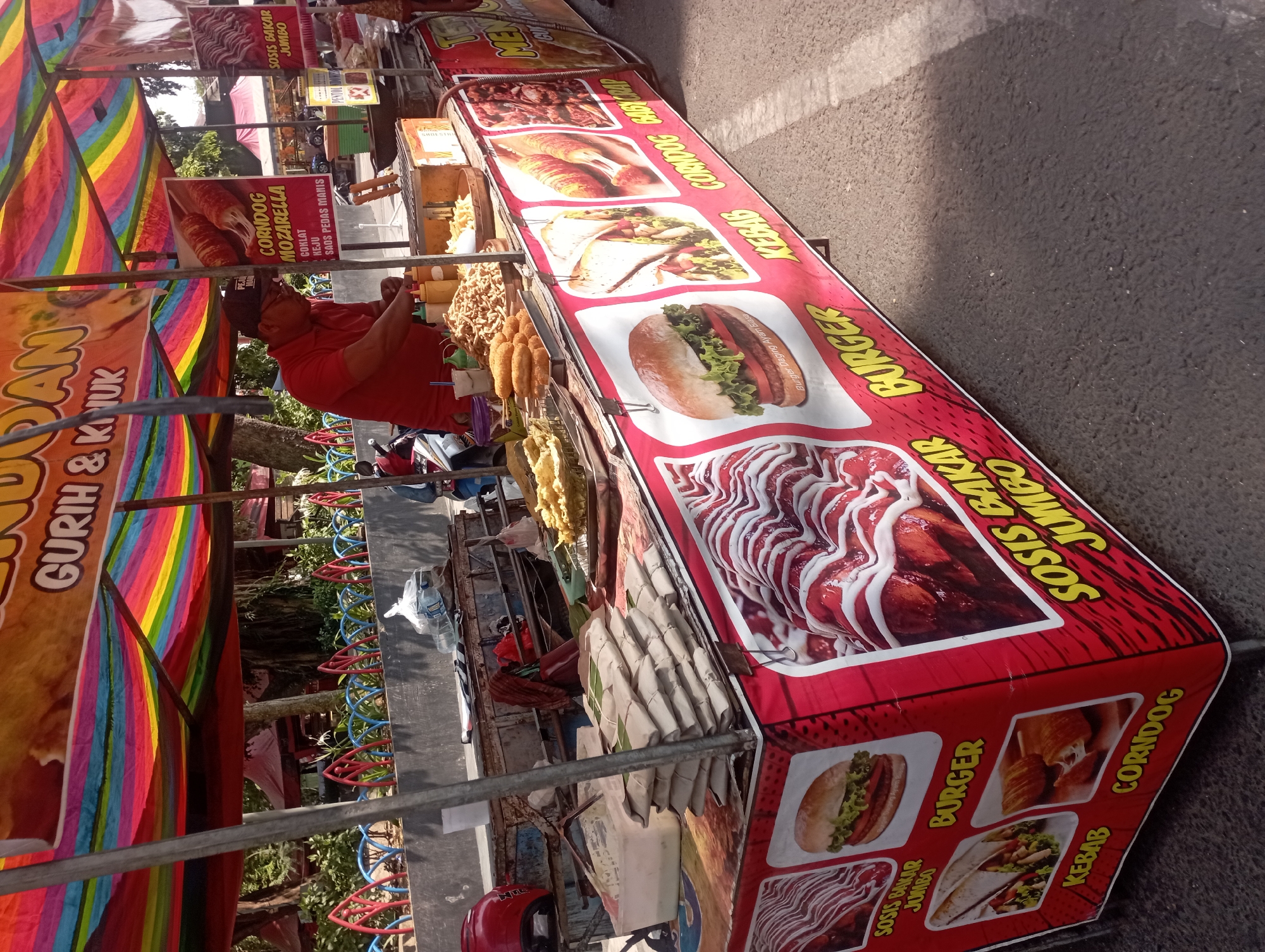A street food vendor is preparing food at a cart in Nganjuk, Indonesia. The cart is covered with a colorful striped awning and adorned with images of various food items like burgers, kebabs, sausages, and corndogs. A man in a red shirt and black cap is seen behind the cart, attending to the food on a grill. Various food items, including fried snacks and bottles of water, are visible on the cart. The background shows trees, a fence, and other stalls, suggesting a public area or market. The ground is paved, and sunlight casts shadows on the scene.