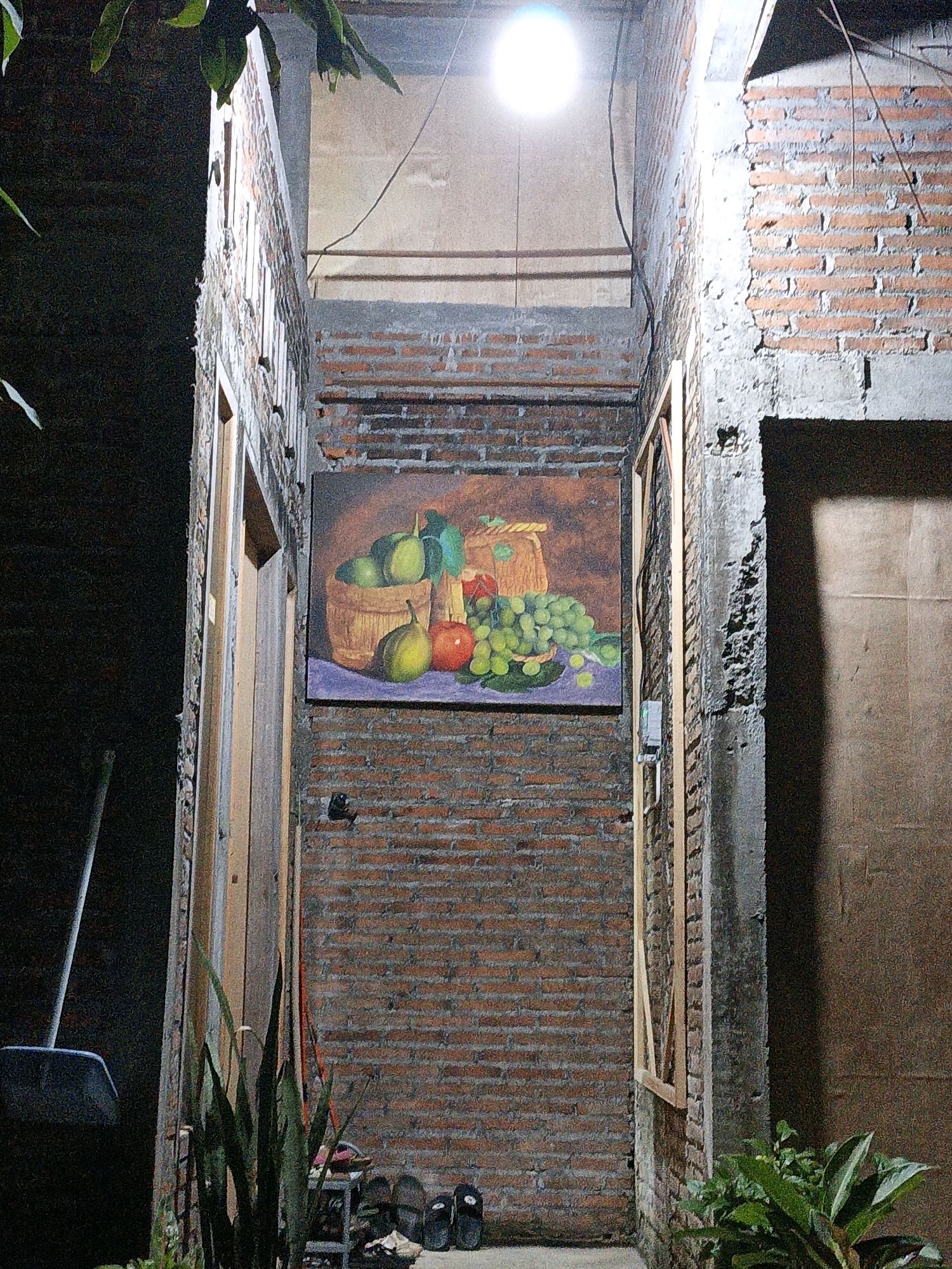 The image depicts an outdoor, narrow corridor between two brick walls at night, illuminated by a bright overhead light fixture. A large painting of a fruit still life, featuring grapes, apples, and pears in a basket, hangs on the left brick wall. To the left of the painting, a wooden door frame is visible, with a wooden door slightly ajar. At the base of the wall, near the door, is a dark broom with a blue dustpan. Further down the corridor, a small rack holds several pairs of sandals and other items. On the right wall, a rough concrete surface is exposed, contrasting with the brickwork. In the foreground, on the left, is a leafy green plant, and on the right, another cluster of green plants. The lighting suggests the image was taken in Cikal, Indonesia, given the architectural style and the presence of common household items. There are no people or animals visible in the scene.